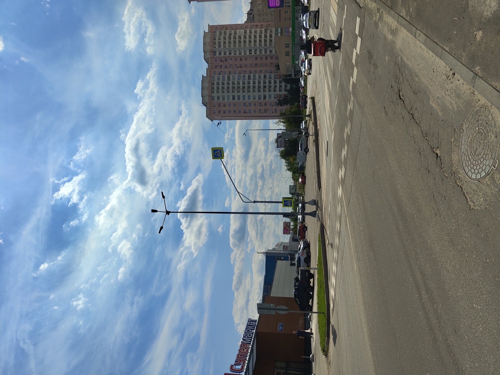 The image shows a wide asphalt road in the city of Zhukovsky, Russia, with a pedestrian crossing and traffic lanes. On the left side of the road, a modern supermarket building with a large red and white sign is visible, along with a parking lot containing several vehicles. Across the street, a tall, pink residential building with many windows stands prominently, indicating an urban setting. Other cars and a scooter with a red delivery box are navigating the intersection. The sky is bright blue with scattered white clouds, suggesting fair weather. A lamppost with multiple lights is positioned on the left side of the road, and a pedestrian crossing sign is visible on the right. The road surface shows some wear and tear, with cracks and uneven patches.
