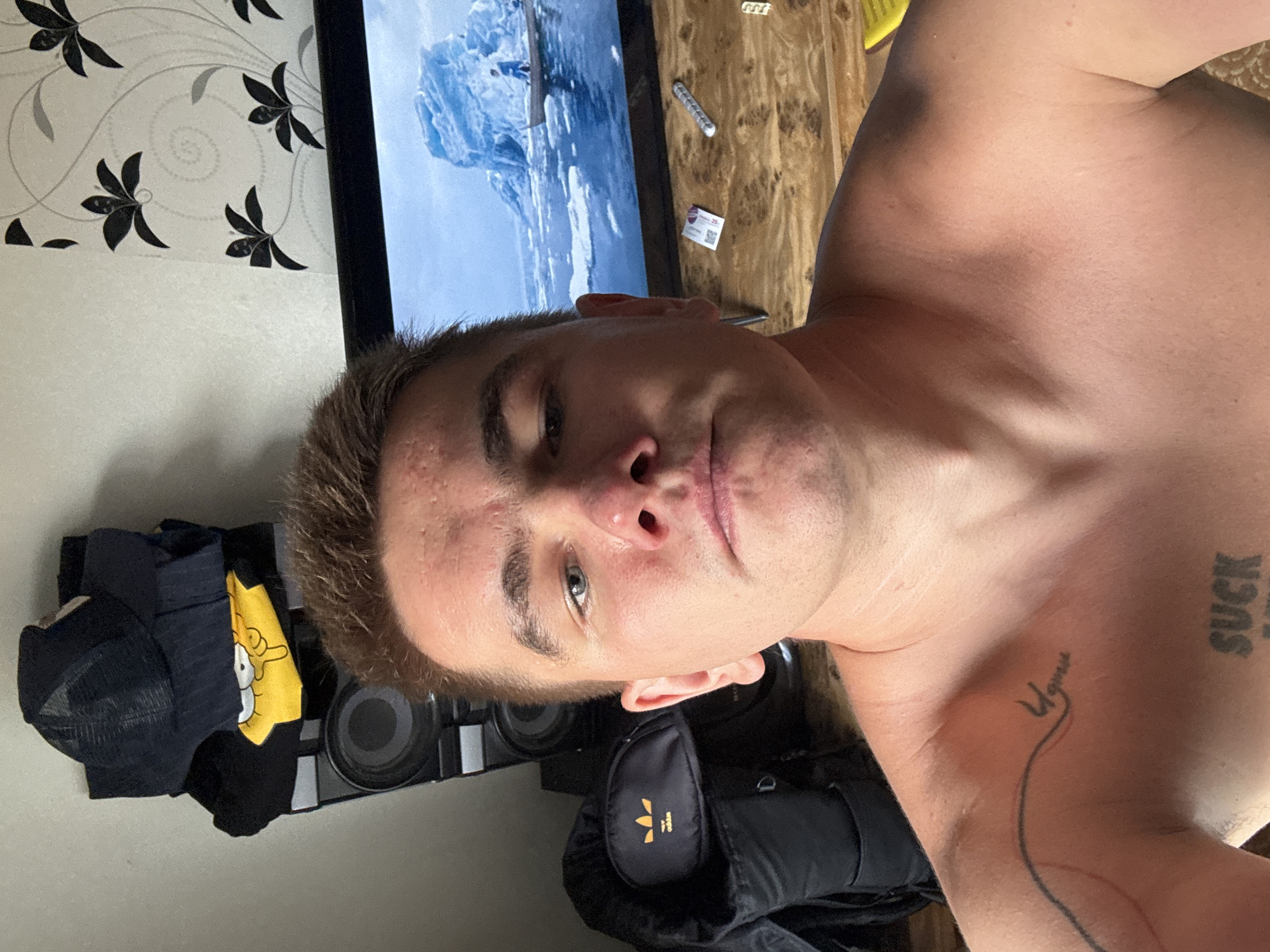 The image is a selfie taken indoors, possibly in a bedroom or living area. A young man is shirtless and looking directly at the camera. He has light brown hair and fair skin with some blemishes on his forehead and cheeks. He is wearing a necklace.
In the background, there are several items visible:
* On the left, a black trucker hat is placed on top of stacked clothing. Below the hat is a black t-shirt with a yellow graphic of Bart Simpson. Underneath the t-shirt are two large speakers stacked vertically.
* To the left of the man's head, a black backpack with an Adidas logo is visible.
* Behind the man and slightly to his right, a television screen displays an image of an icy landscape with a small boat and two figures.
* On the wooden surface below the television, there are a few small items: a white card with a QR code, a silver cylindrical object, and a yellow object, possibly a lighter.
* The wall behind the television has patterned wallpaper with black floral designs.
* The man has two tattoos visible on his upper body: one on his left shoulder area that appears to be cursive text, and another on his chest that reads "SUCK".
The overall impression is an informal, personal snapshot. The location in the city of Shcherbinka, Russia, is not directly indicated by any visual cues within the image itself.