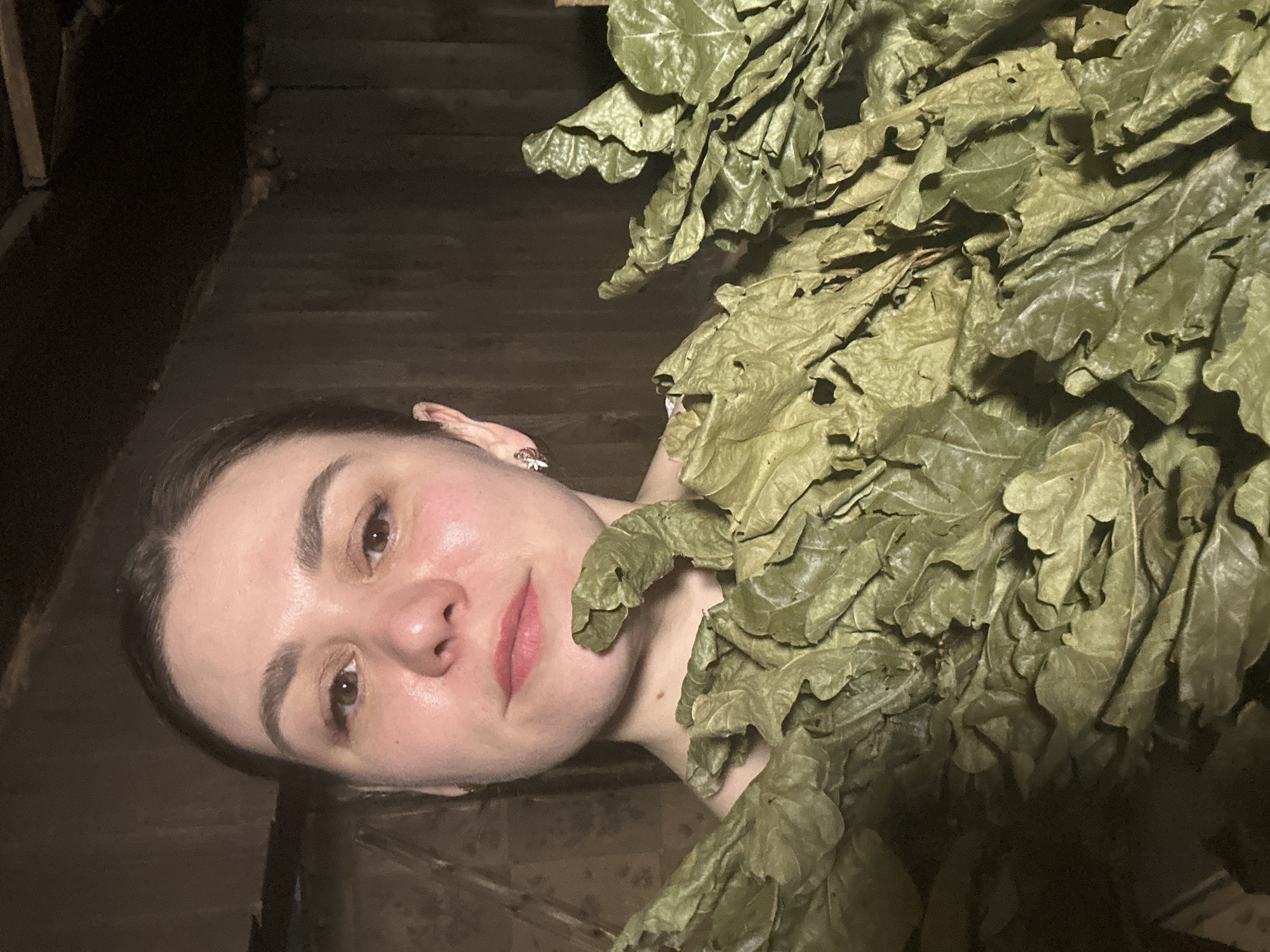 A woman is in a sauna or bathhouse in Chaikovsky, Russia. She is partially covered by oak leaves, possibly a venik used for massage in Russian bathhouses. The background appears to be wooden walls and flooring.
