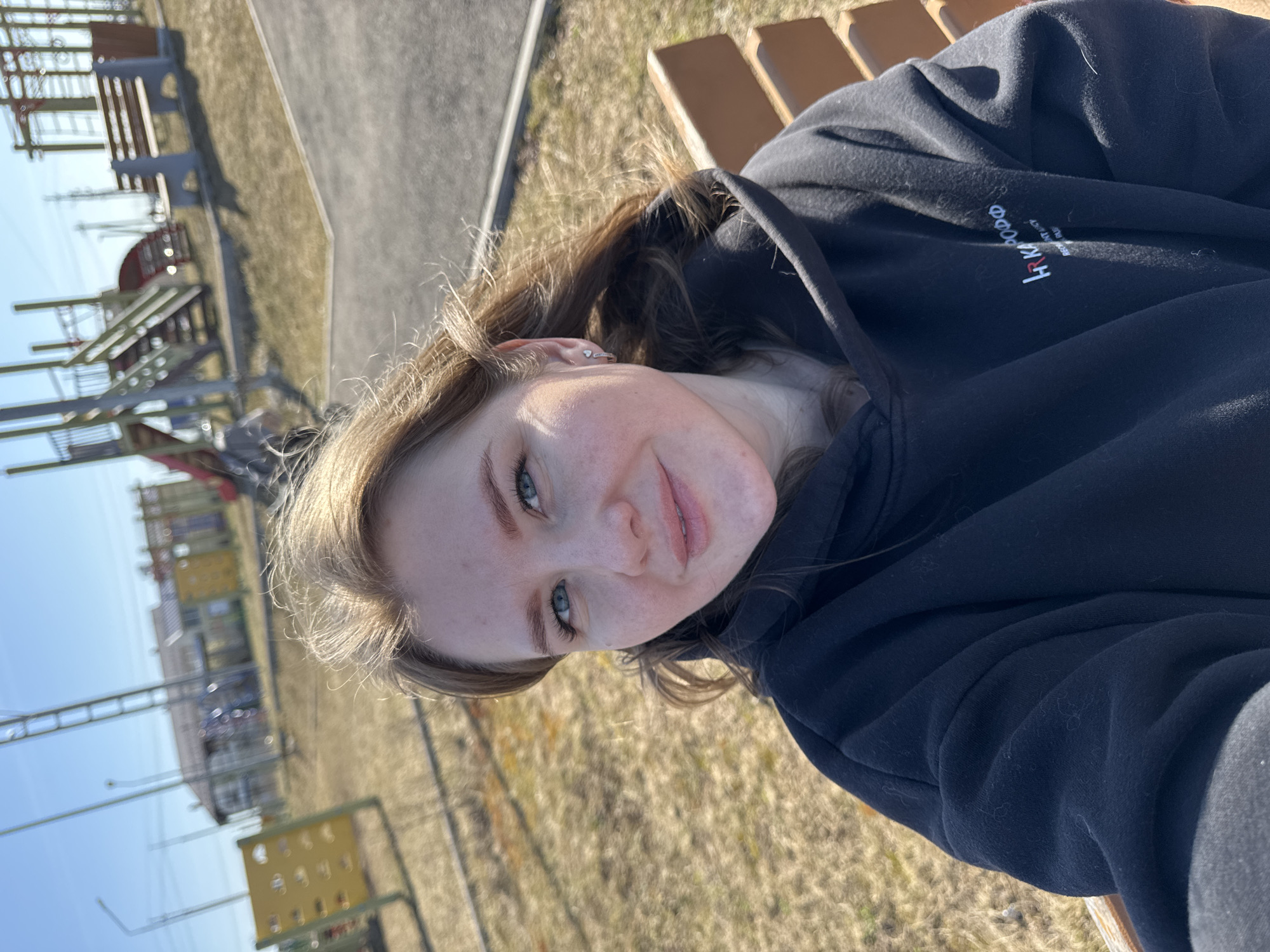 The image is a selfie of a young woman with fair skin, light brown hair, and blue eyes. She is wearing a black hooded sweatshirt. She is sitting on a light brown bench. The background appears to be a playground with metal structures and a pathway. The location is Горный, Russia.
