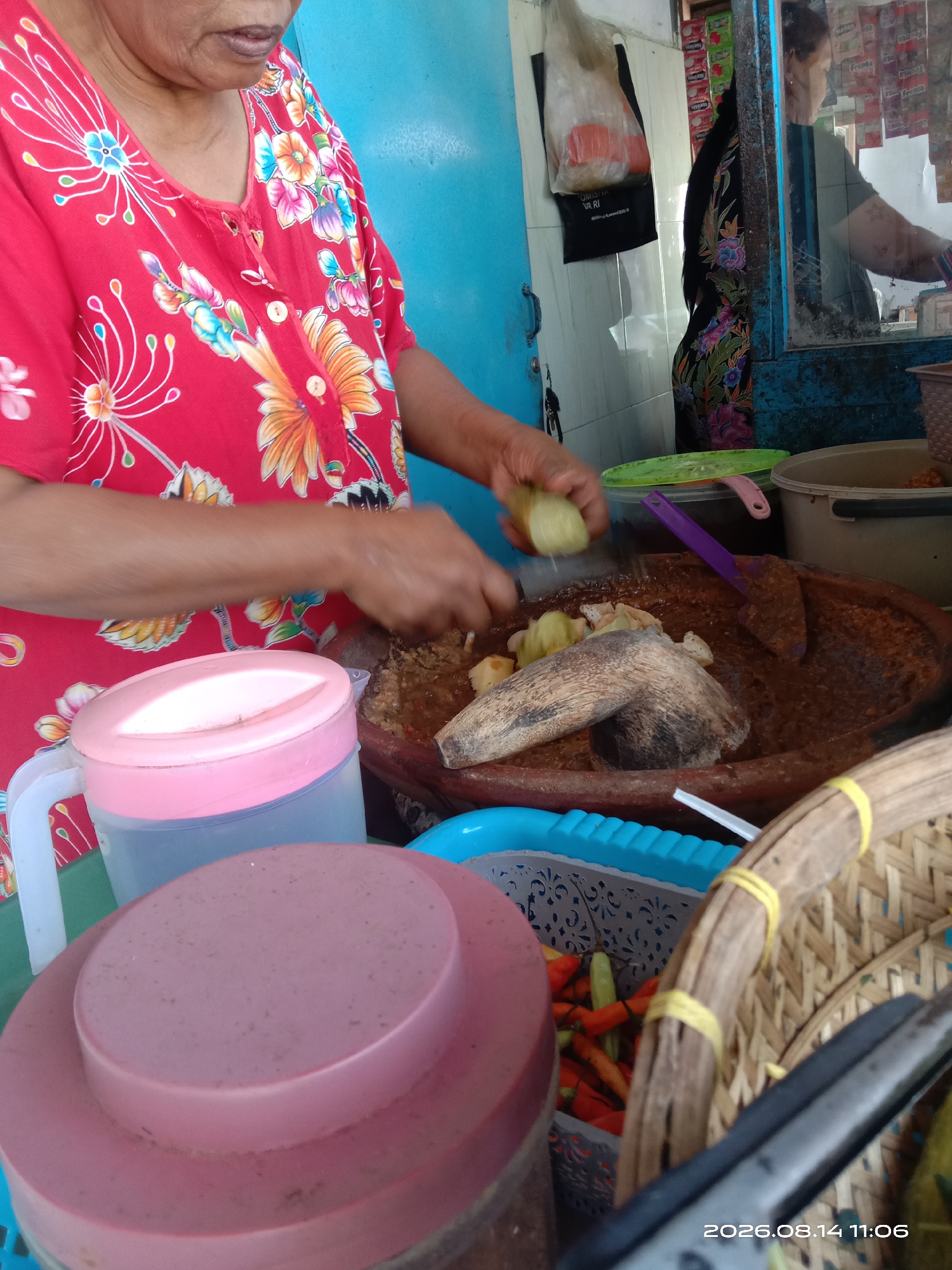 In Blimbing, Indonesia, a person wearing a vibrant red, floral-patterned shirt is preparing food. They are using a mortar and pestle, made of wood and stone respectively, to mash ingredients in a large, brown, oval-shaped bowl. Various food items and containers are visible in the scene. To the left of the mortar is a pink-lidded pitcher. In front of the pitcher, another pink lidded container is visible. To the right of the mortar, a blue basket contains small chilies, and a woven basket sits nearby. In the background, a blurry reflection in a window shows another person. The environment appears to be an outdoor food stall or a small shop, with a blue wall as a backdrop. A date stamp in the lower right corner reads "2026.08.14 11:06".