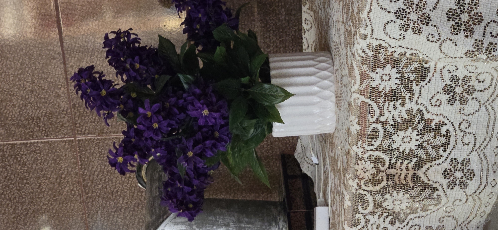 An artificial floral arrangement of purple flowers with green leaves sits in a white patterned vase on a white lace-like tablecloth. Behind the arrangement is a textured, copper-colored wall. To the left of the vase, a portion of a cylindrical object, possibly metal, is visible. A white rectangular object and a white cord are also on the tablecloth. The image appears to be taken indoors, likely in a residential setting. There is no information about the location in Tel, Azerbaijan in the provided image.