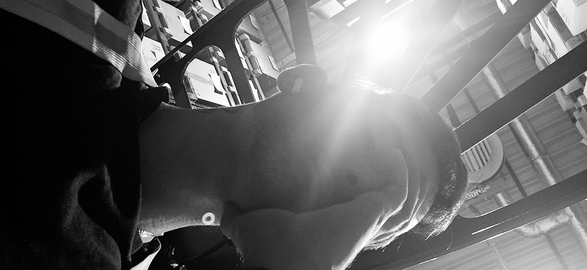 A high-contrast, black and white image, taken from a low-angle perspective, captures an individual looking upwards within an indoor industrial setting. The subject, seen from the neck and shoulder, occupies the lower-left portion of the frame. An earbud is visible in their ear, and they are wearing a dark garment with a subtle striped pattern on the shoulder. A small, circular white object, possibly a button or sensor, is present on their neck.
The background reveals a warehouse or large storage facility in Plovdiv, Bulgaria. Prominent dark structural beams or metal shelving units cut across the upper frame. Behind these, numerous cardboard boxes are stacked on shelves, indicating storage. A strong, bright light source in the upper right quadrant creates significant lens flare and highlights, suggesting either a window with natural light or a powerful overhead lamp. Parts of the ceiling structure, including what appears to be a large circular ventilation fan and ductwork, are also visible. The high contrast emphasizes the textures and shapes within the scene. Faint, indistinct text resembling "BBBB" and "EEE" along with barcodes can be seen on some distant boxes. The activity appears to be the individual observing their surroundings from a low vantage point, possibly while working or resting in this industrial environment.