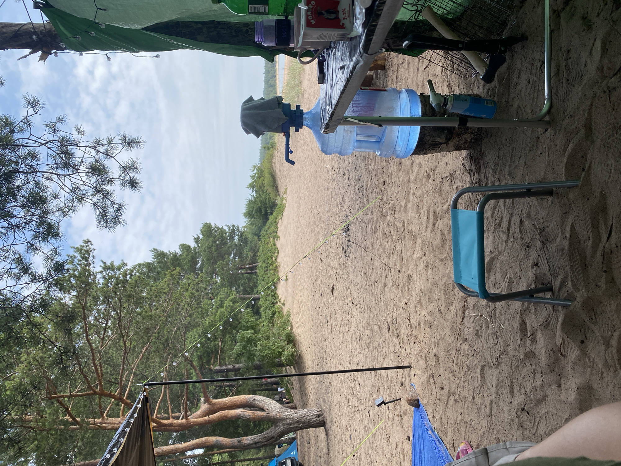 This is a medium shot of a campsite on a sandy beach with pine trees in the background. The campsite is set up under a green tarp, and there is a large blue water jug with a pump on top. A small blue chair sits on the sand in front of the water jug. A table is set up next to the water jug, with a cup, a can of condensed milk, and other items on it. In the background, there are tents and a car. The sky is cloudy. The location appears to be in or near Baltiiskoe, Russia, given the presence of pine trees and a beach setting that is consistent with coastal regions in Russia. There are no people clearly visible in the image, only partially visible legs in the foreground. The scene depicts a temporary outdoor living space, likely for camping or recreational activities.