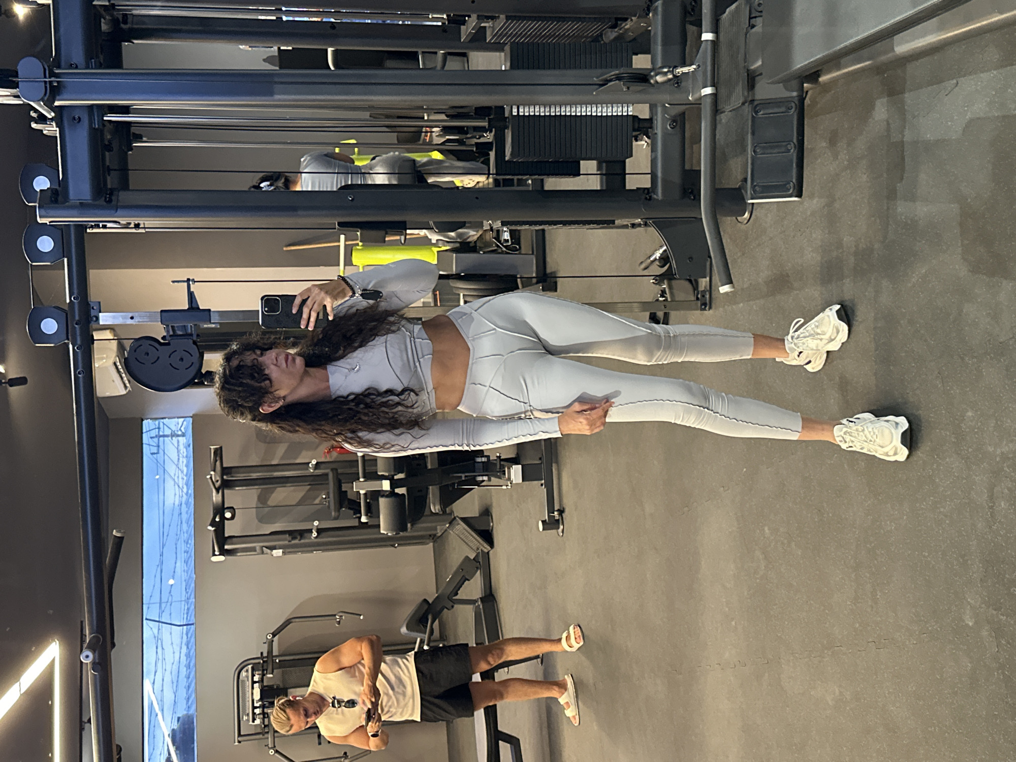 In a gym located in Kuta Selatan, Indonesia, a woman is taking a selfie in the mirror. She is wearing a light gray long-sleeved crop top and matching leggings, with white sneakers. Her dark, curly hair is down, and she is holding her phone to take the picture. In the background, a man is standing near a weight machine, looking at his phone. He is wearing a light-colored sleeveless shirt, black shorts, and white sandals. The gym has a gray floor and is equipped with various weight machines.