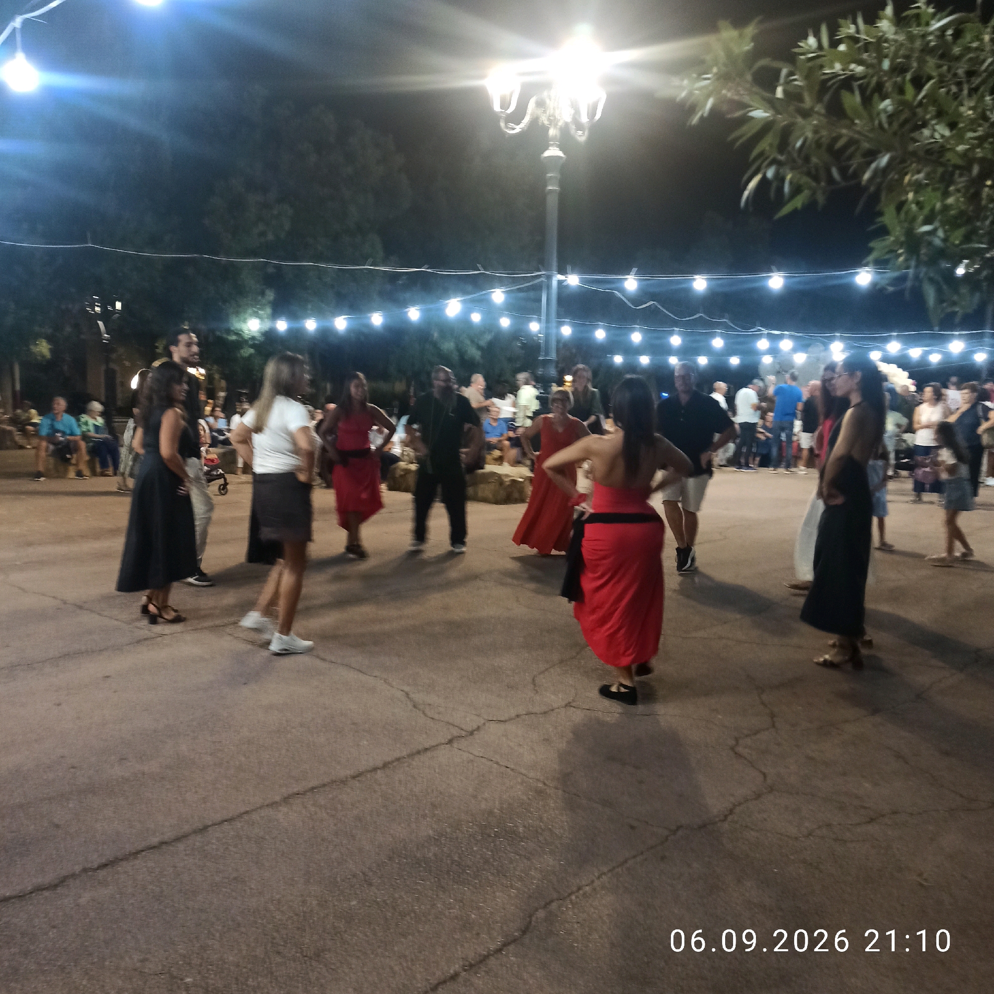The image depicts an outdoor evening gathering with a group of people in what appears to be a public square or park. A street lamp is visible in the background, along with strings of decorative lights. Several individuals are present, some of whom appear to be dancing or socializing. The ground is paved with stone or concrete. In the lower right corner, a timestamp is visible: "06.09.2026 21:10". The scene is illuminated by artificial lighting, creating a festive atmosphere. The presence of a date stamp suggests this image was taken on September 6, 2026, at 9:10 PM.