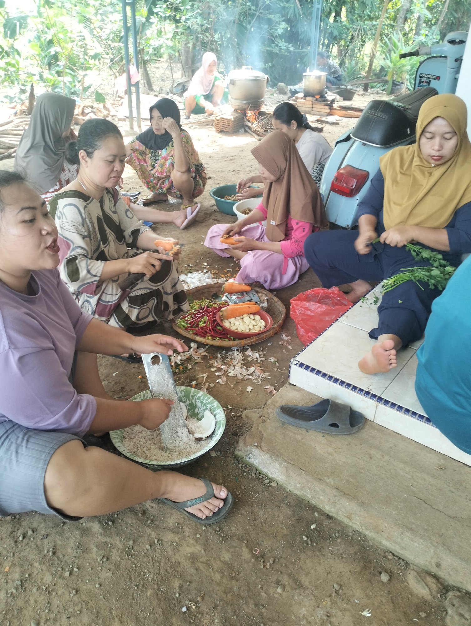 In Kaligarang, Indonesia, a group of women are gathered outdoors, seemingly preparing food. The scene is informal, with some sitting on the ground and others on a low concrete ledge. In the foreground, a woman in a purple t-shirt and grey shorts is grating coconut into a green bowl with a metal grater. To her right, another woman in a patterned dress and headscarf is also preparing food, with carrots and other vegetables on a woven mat.
Behind them, several other women are engaged in similar activities. One woman, wearing a yellow hijab and blue clothing, is holding a bunch of fresh green herbs. Another woman in a brown hijab and pink shirt is peeling a carrot. Further back, two women are tending to a cooking fire where pots are steaming, indicating that cooking is in progress.
The environment appears to be rural, with lush green vegetation in the background and a dirt ground surface. An old light blue scooter is parked to the right of the scene, suggesting a mode of transportation. The overall atmosphere is one of community and shared activity, likely a traditional gathering or event.