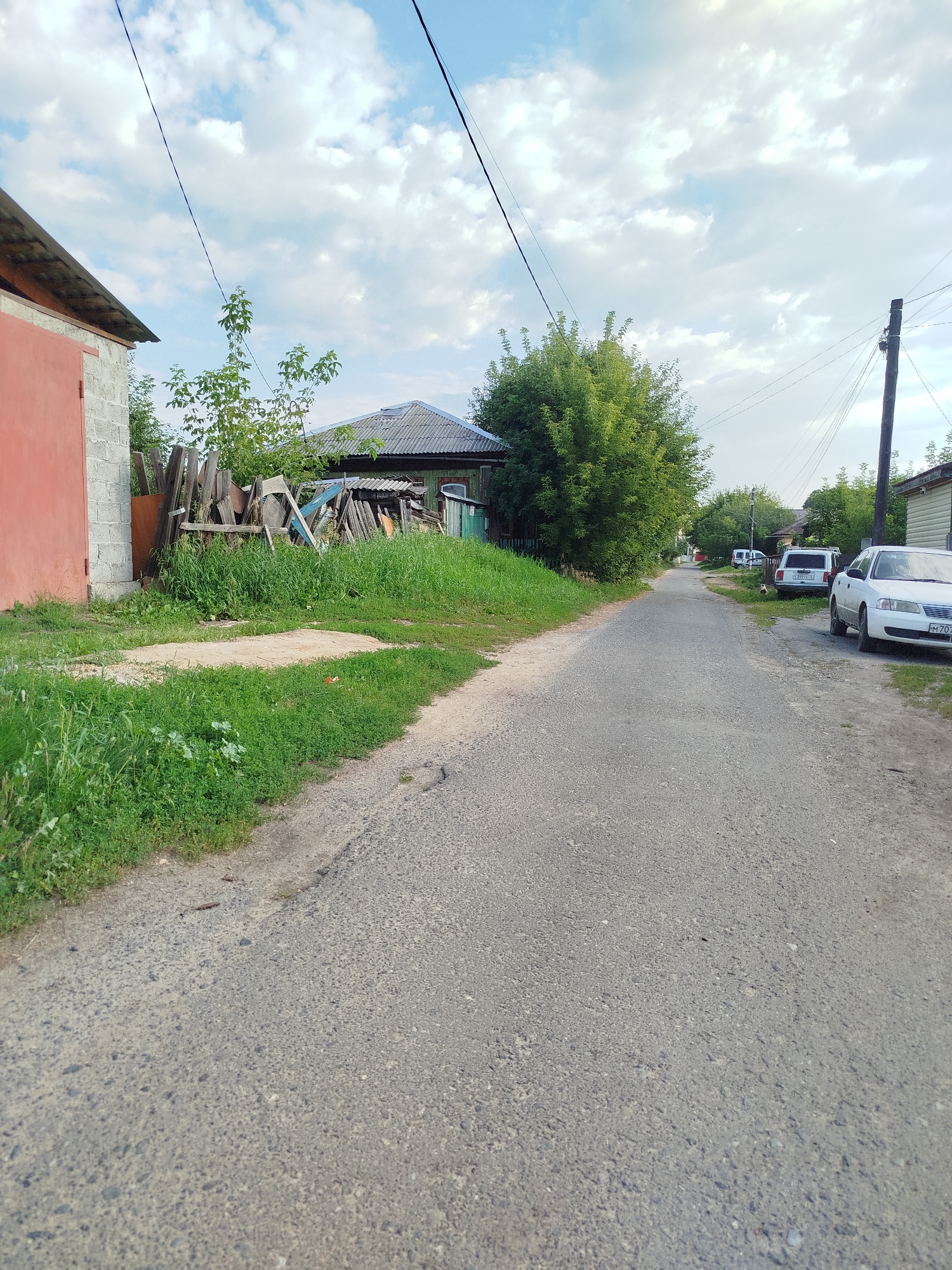 The image shows a rural road in Zavodoukovsk, Russia. On the left, there is a building with a red door and a grey block wall, adjacent to a pile of wooden debris and overgrown grass. Further down the road, there are several parked cars, including a white sedan and a white minivan. Trees and houses line the right side of the road, with power lines stretching across the sky. The road surface appears to be asphalt with some cracks and patches. The sky is cloudy with patches of blue.