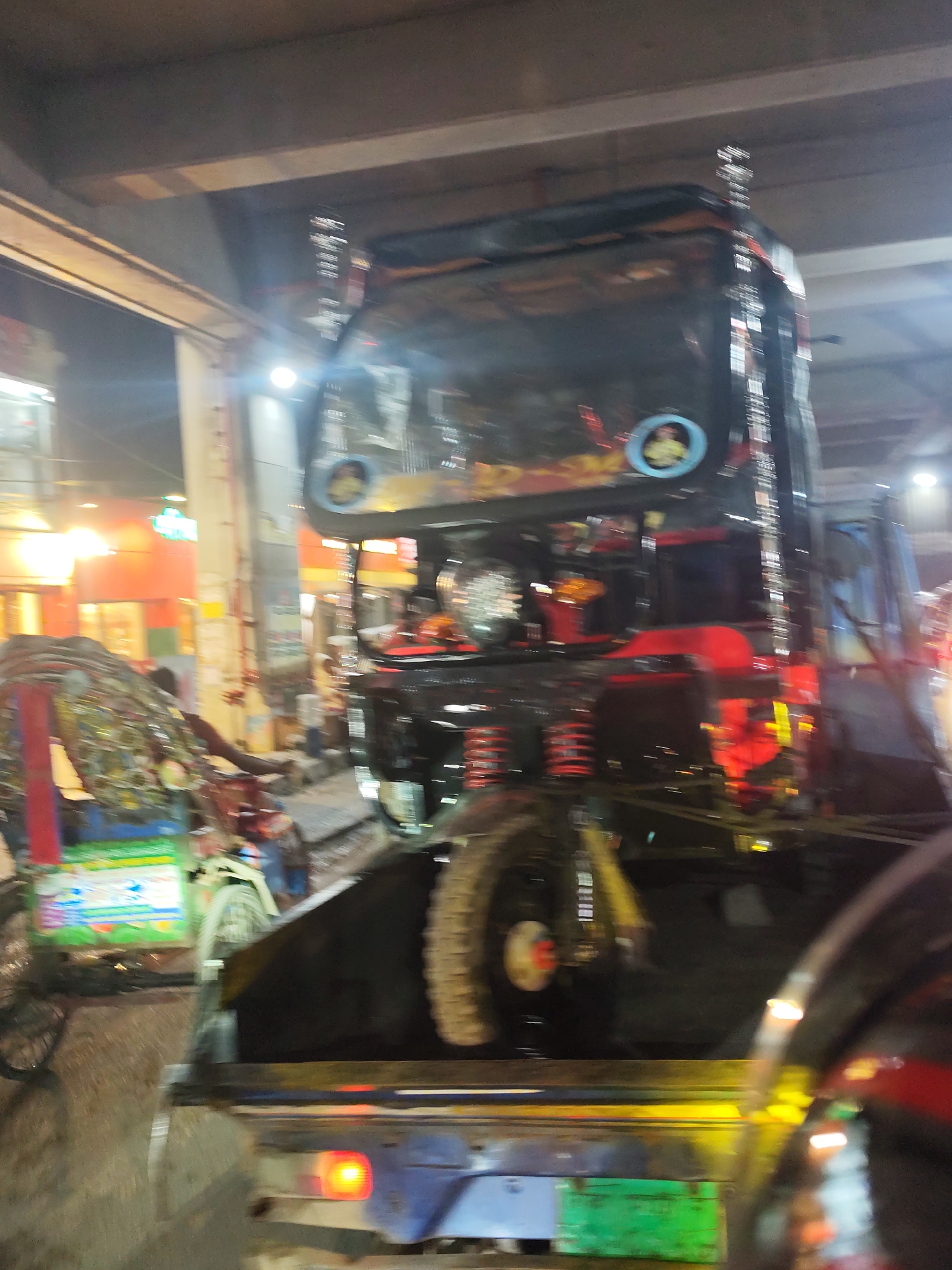 The image captures a nighttime street scene in Dhaka, Bangladesh. A decorated auto rickshaw, commonly known as a CNG, is prominently featured, with its front and back partially visible. The CNG displays extensive chrome detailing and decorative elements. To the left, a colorful cycle rickshaw is partially visible. The scene is illuminated by artificial lighting, including streetlights and vehicle lights, creating a bright environment. The location appears to be under a bridge or overpass in a busy urban area. Other vehicles and possibly people are in the background.