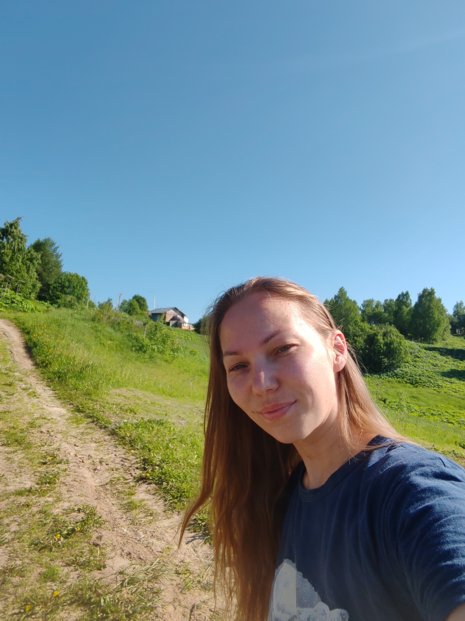 A young adult female, with long light-brown hair and a subtle smile, is captured in a selfie in Чухлэм, Russia. She wears a dark blue t-shirt with a partially visible light-colored graphic print. Her face exhibits faint freckles. The background reveals a bright, clear blue sky above a verdant, grassy hillside. A dirt path ascends from the lower-left, curving uphill. Deciduous trees with rich green foliage dot the landscape, particularly along the left side of the path and across the upper right of the hill. A rustic wooden house with a dark roof stands on the hillside in the distance, partially concealed by greenery. Strong sunlight illuminates the scene, indicating a clear day during spring or summer.