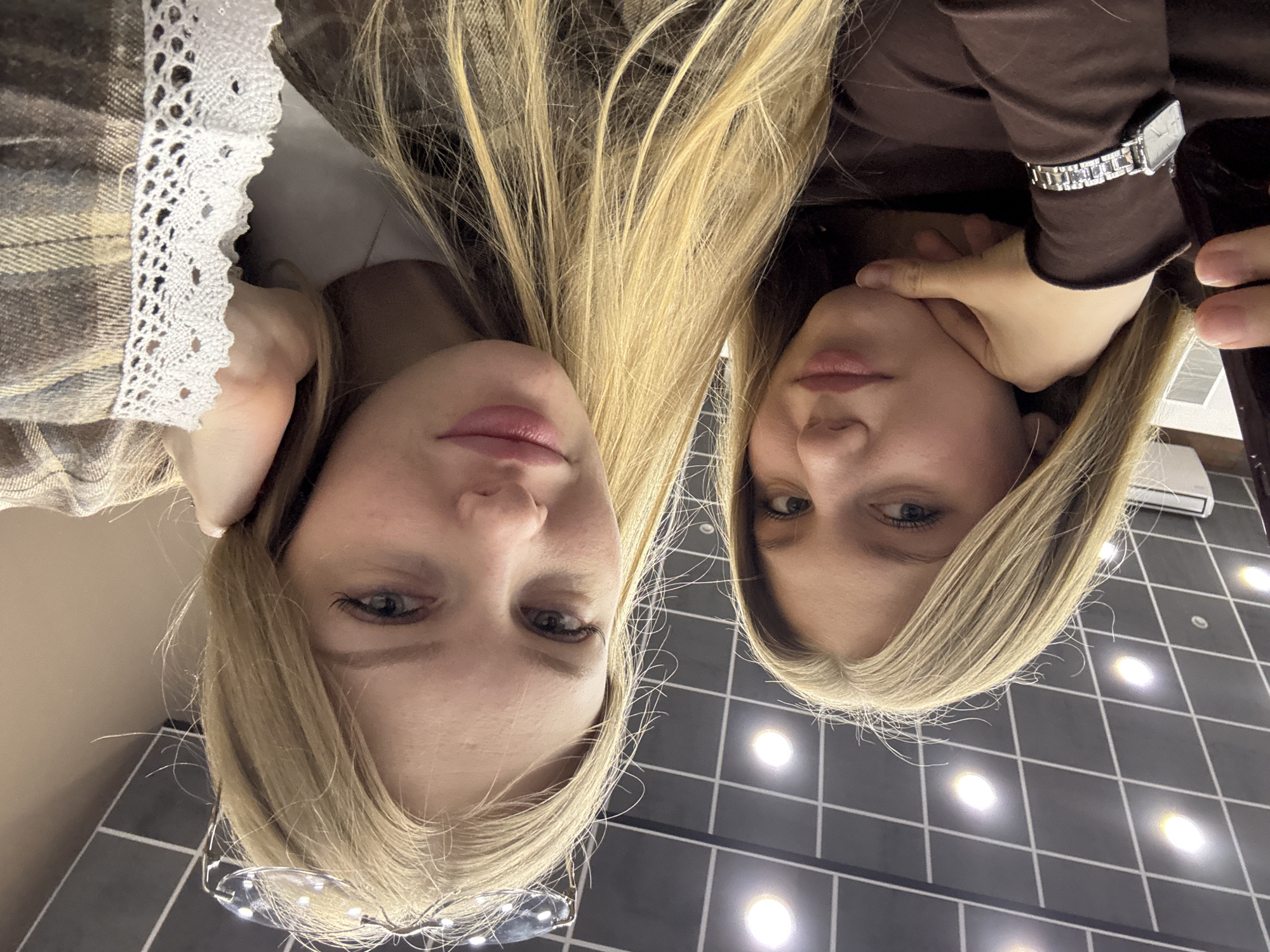 This image is a close-up, low-angle portrait of two young women with long, straight blonde hair, presented upside down. When oriented correctly, both subjects are looking directly downwards towards the camera with neutral to slightly smiling expressions.
The woman on the left is partially obscured by her hair but shows a portion of a plaid top, likely in brown and beige tones, with decorative white lace trim along a collar or cuff. A pair of clear-framed glasses rests on her nose.
The woman on the right wears a solid dark brown long-sleeved top. A silver-toned wristwatch with a square face is visible on her left wrist, and her right hand is gently touching her chin or jawline.
Their blonde hair cascades downwards, almost meeting in the center of the frame. The background consists of a modern indoor ceiling, tiled with dark grey or black squares and illuminated by numerous bright, circular recessed light fixtures. This suggests an indoor commercial or public setting.
The image appears to be a casual selfie or portrait. No specific activity beyond posing for the camera is evident. There is no visible text within the frame. The location context provided is Perm, Russia.