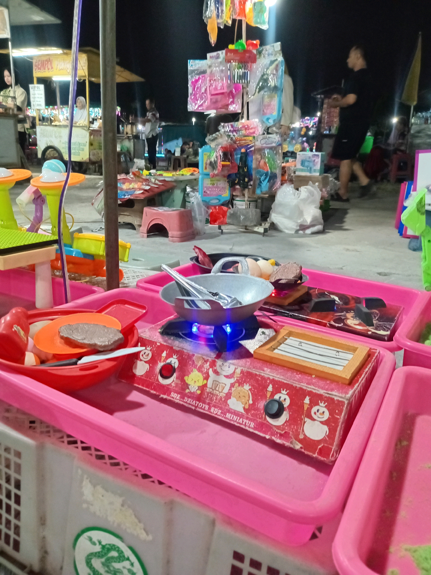 The image depicts an outdoor night market scene in Sedati Agung, Indonesia. In the foreground, a toy stove with a blue flame is set atop a red box adorned with cartoon snowmen and text that reads "SDZ... MINIATUR". A toy frying pan with a piece of brown "food" and utensils sits on the stove. This toy cooking set is placed within a pink plastic tray, which rests on top of a white plastic basket with a checkered pattern. To the left, a red plastic bowl contains various toy kitchen items, including orange plates and a knife. In the mid-ground, more toy stands and carts are visible, displaying an assortment of colorful toys and small packaged goods. Several people are present, some appearing to be vendors and others customers browsing the stalls. The overall environment is dimly lit, with artificial lights illuminating the market area. The background shows more stalls and a dark night sky.