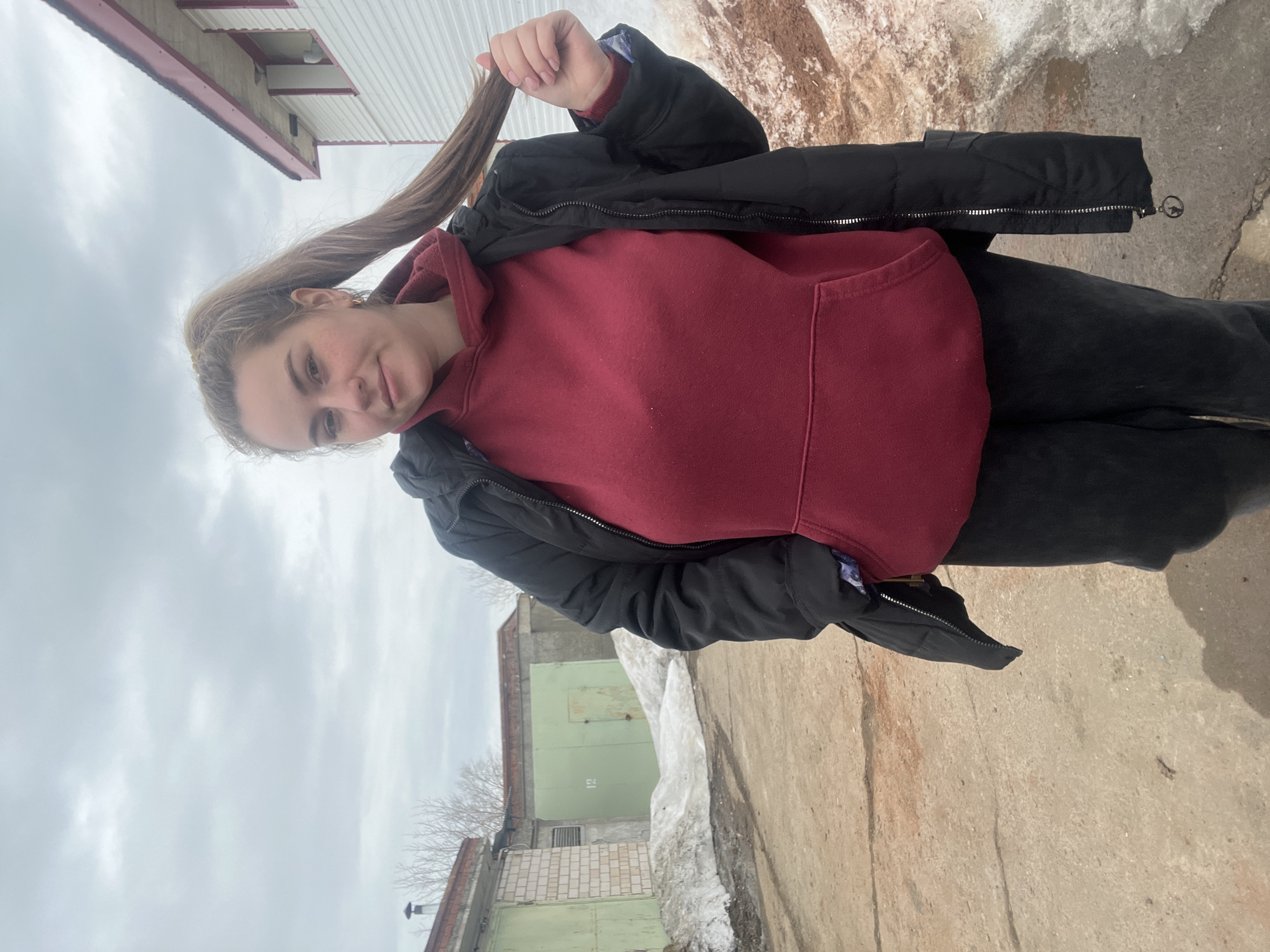 A young woman stands outdoors in Chaikovsky, Russia. She is wearing a dark jacket over a red hoodie and dark pants, holding a strand of her light brown hair in her right hand. The background includes buildings, possibly garages, some with numbers visible. There is melting snow and bare trees suggesting a late winter or early spring day. The sky is overcast. The surface she is standing on appears to be concrete or asphalt.