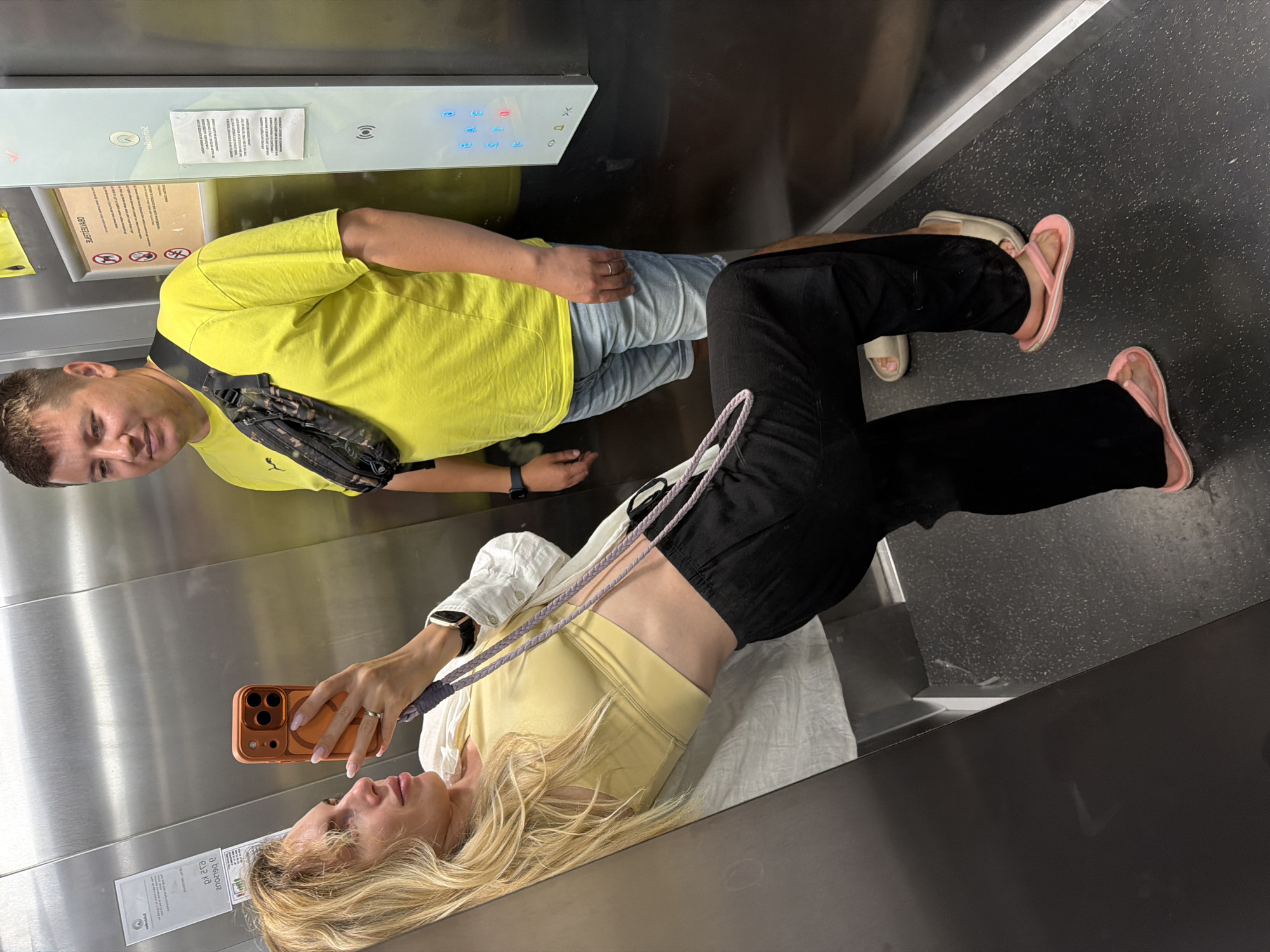 In an elevator in Budva, Montenegro, a woman with blonde hair takes a selfie with her phone. She is wearing a yellow sports bra, black pants, and pink flip-flops. Leaning against the elevator wall, she holds a purple strap in her hand. A man stands behind her, looking towards the camera. He wears a bright yellow t-shirt, light blue denim shorts, and a camouflage fanny pack across his chest. He also wears a ring on his left hand and a watch on his left wrist. The elevator's interior features metallic walls, a control panel with illuminated buttons, and floor markings. The floor is dark gray with a speckled texture.