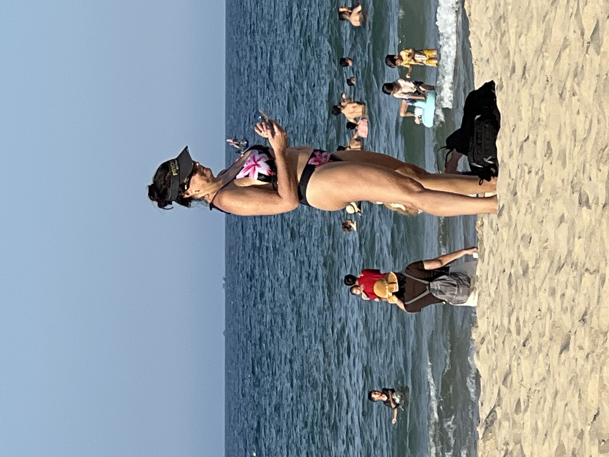 The image depicts a scene at My Khe Beach in Da Nang, Vietnam. In the foreground, a woman in a floral bikini stands on the sandy shore, holding a smartphone. She is wearing a black visor. Behind her, the ocean is populated with various people enjoying the water. Several children are visible, some playing near the shore on a blue inflatable. Other individuals are swimming or wading in the sea. The sky is clear and blue, with a calm ocean surface. The overall impression is a typical day at a crowded beach.