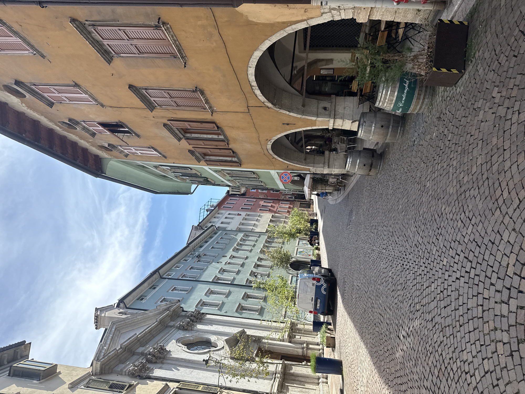 The image captures a narrow, cobblestone street in Trento, Italy, likely a historic district. The street is lined with multi-story buildings displaying distinct architectural styles. On the left, a light-colored building with a classical facade and an arched entrance is visible, possibly a church or public building, with lampposts and trees lining the walkway. Adjacent to it, a row of pastel-colored buildings stretches into the distance, featuring numerous windows and balconies. The right side of the street is dominated by a building with an ochre-colored facade, characterized by arched porticoes at the ground level, creating shaded walkways. Several wooden barrels are placed along the portico, one bearing green lettering. Outdoor seating for what appears to be a cafe or restaurant is visible under the porticoes. A dark blue van is parked on the left side of the street. The sky above is blue with wispy white clouds. The scene is well-lit by sunlight, casting shadows on the cobblestones.