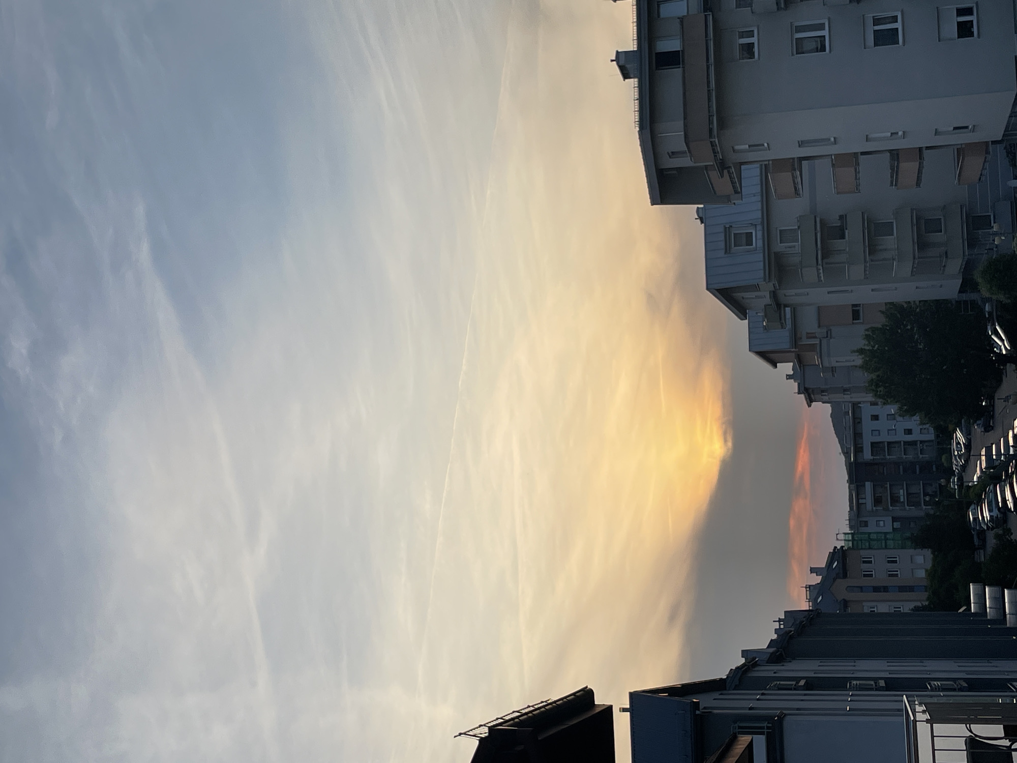 The image captures an urban or suburban landscape under a vivid sky during either sunrise or sunset. The sky dominates the upper and left portions of the frame, exhibiting a striking gradient from light, hazy blue on the left to intense yellow, orange, and reddish hues towards the right, suggesting the low position of the sun. Wispy, elongated clouds or contrails stretch across the sky, particularly noticeable in the cooler blue areas and extending into the warmer tones.
Along the right side and across the bottom of the image are several multi-story residential buildings. These modern structures feature numerous windows and balconies, and their facades appear in muted shades of gray, white, and brown, with some sections in shadow or silhouetted against the bright sky. In the immediate foreground, the dark outlines of building rooftops and upper floors are visible at the bottom of the frame.
In the far background, beyond the main cluster of buildings, a gentle slope of hills or mountains is faintly visible, along with some trees. There are no people or discernible activities depicted in the scene. The overall impression is one of peaceful urbanity bathed in the dramatic, warm light of dawn or dusk. No visible text is present.