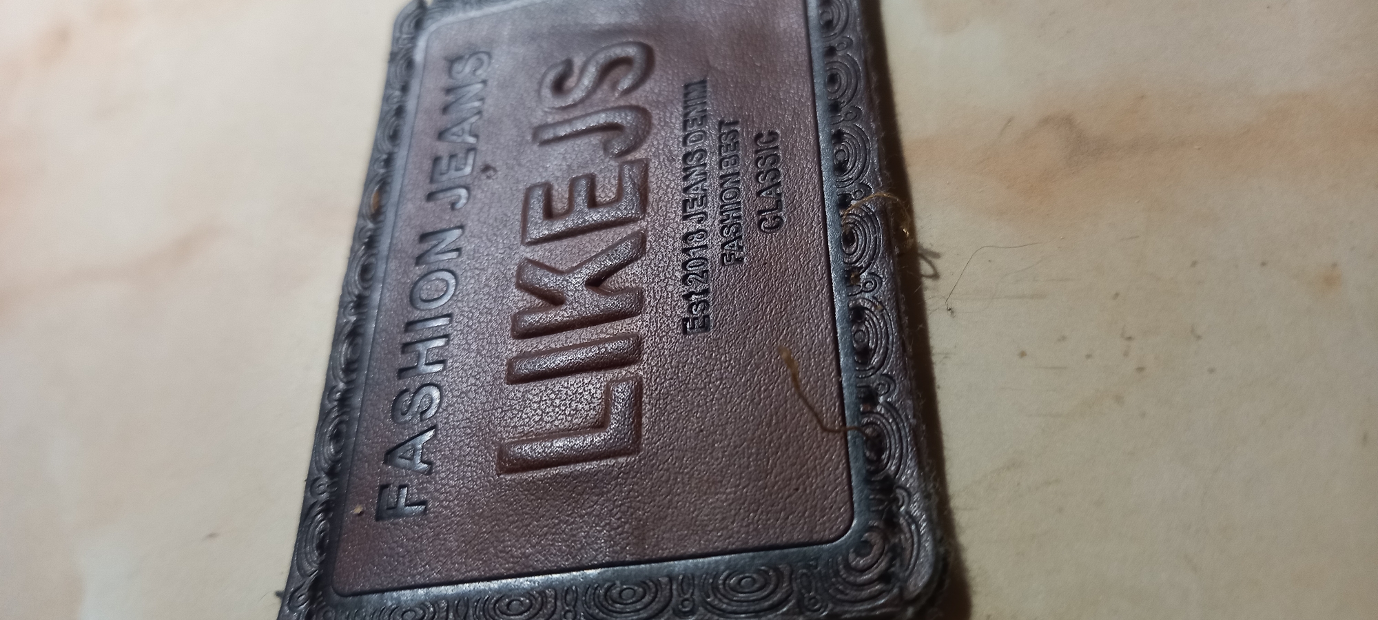 A close-up, angled photograph captures a brown, rectangular patch, likely made of leather or a synthetic material, embossed with text and framed by a dark, ornate border. The central, largest embossed text reads "LIKEJS," with "FASHION JEANS" positioned above it. Below "LIKEJS," three lines of smaller embossed text state "Est 2013 JEANS DENIM," "FASHION BEST," and "CLASSIC." The brown patch displays a pebbled texture, contrasting with the smooth, darker, relief-patterned border featuring intricate scrollwork. The object is situated on a light-colored, possibly beige or off-white, surface that exhibits minor discolorations, specks, and fine fibers. The scene is located in Kryvyi Rih, Ukraine. No individuals, actions, or direct interactions are visible.