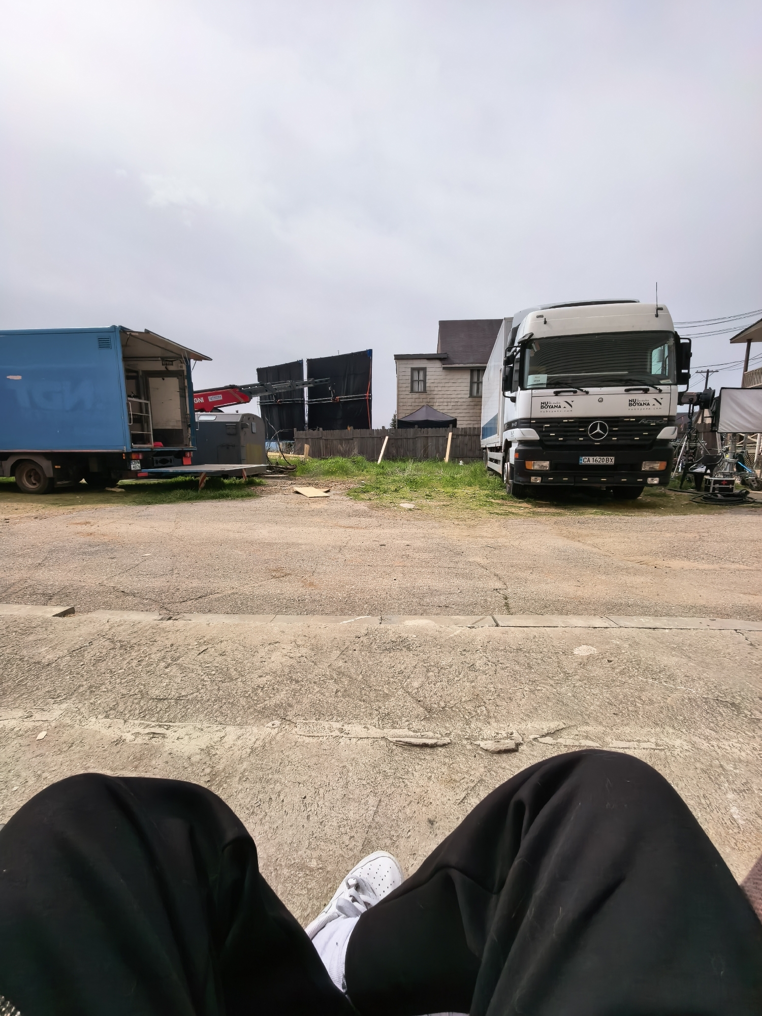 The image presents a scene in Dolna Malina, Bulgaria, characterized by a staging area with logistical vehicles likely used for a film or video production.
Visible are two large trucks: a blue one with a partially open back, and a white Mercedes-Benz truck with visible "NU Boyana" branding, indicating its affiliation with the film studio. A red crane is visible near the blue truck. Behind the vehicles and a wooden fence, there is a backdrop structure of black screens. To the right of the white truck is a residential structure, potentially part of the backdrop.
The foreground presents a rough, concrete surface that extends up to where the photographer is positioned. A person's legs, clothed in black sweatpants with a white shoe visible on one foot, suggest the photographer is seated or positioned low to the ground.
The sky is overcast, providing indirect light to the scene. The overall setting suggests an outdoor film set or production site with essential elements for logistical support and scene creation. The presence of a residential structure as a backdrop also points to an on-location shoot in a partially developed or industrial area.