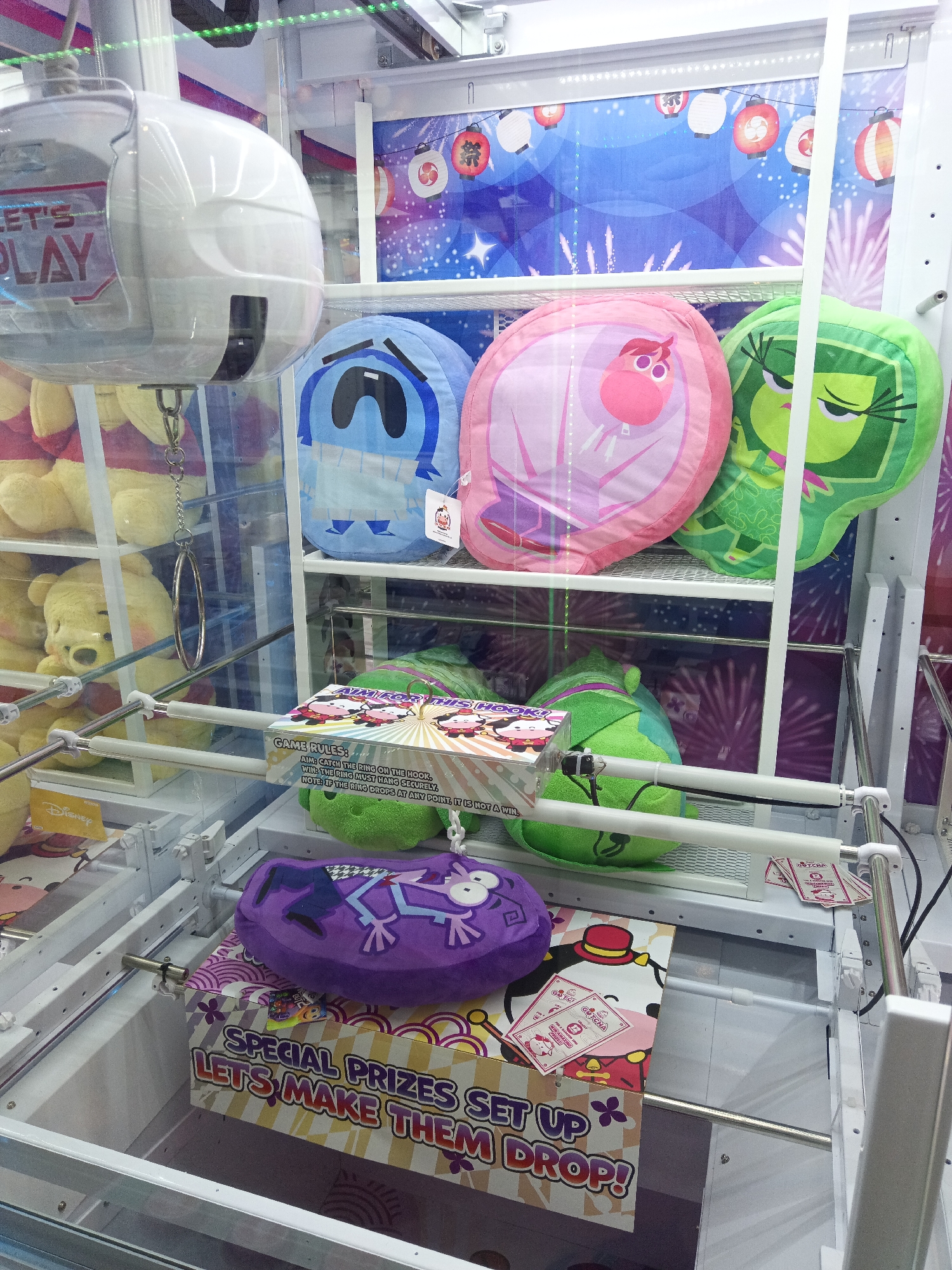 This is a photograph of a claw machine located in Kuala Lumpur, Malaysia, likely within a shopping mall or entertainment center, evidenced by the display of prizes and the typical arcade environment. The machine contains a variety of plush toys, including several character-based ones. On the upper shelves, a blue plush with a sad face and large eyes, a pink plush with a bird-like character, and a green plush with angry eyebrows are prominently displayed. Below these, a small box with "GAME RULES" printed on it is visible, alongside two more green plush toys. In the lower section of the machine, a purple plush toy with a surprised expression and a frog-like character is placed on top of a colorful box featuring cartoon characters and the text "SPECIAL PRIZES SET UP LETS MAKE THEM DROP!". A metal claw mechanism is suspended above the prizes. The background of the machine is decorated with an image of colorful lanterns and fireworks. The overall scene depicts a typical claw machine prize selection.