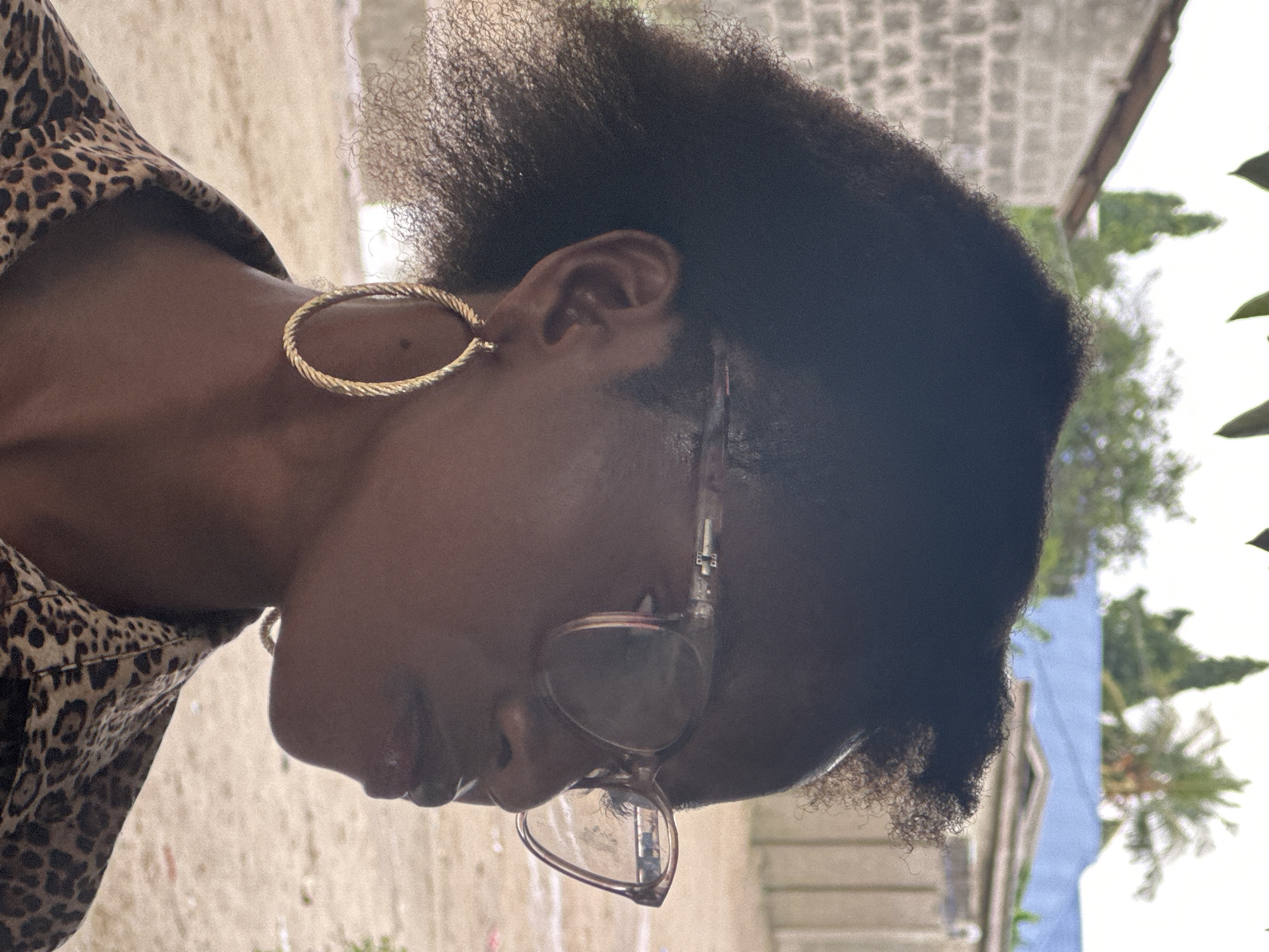 The medium shot image features a Black woman from the chest up, in profile, facing right. She has dark, curly hair, wears large, gold hoop earrings, and clear-rimmed glasses with a rose tint. Her lips are glossy, and she is wearing a leopard-print collared shirt. The background suggests an outdoor setting with a brick building on the left, a palm tree and other foliage in the middle ground, and a pale blue structure and buildings in the distance, characteristic of the city of Badagry, Nigeria. The ground appears sandy or dirt.