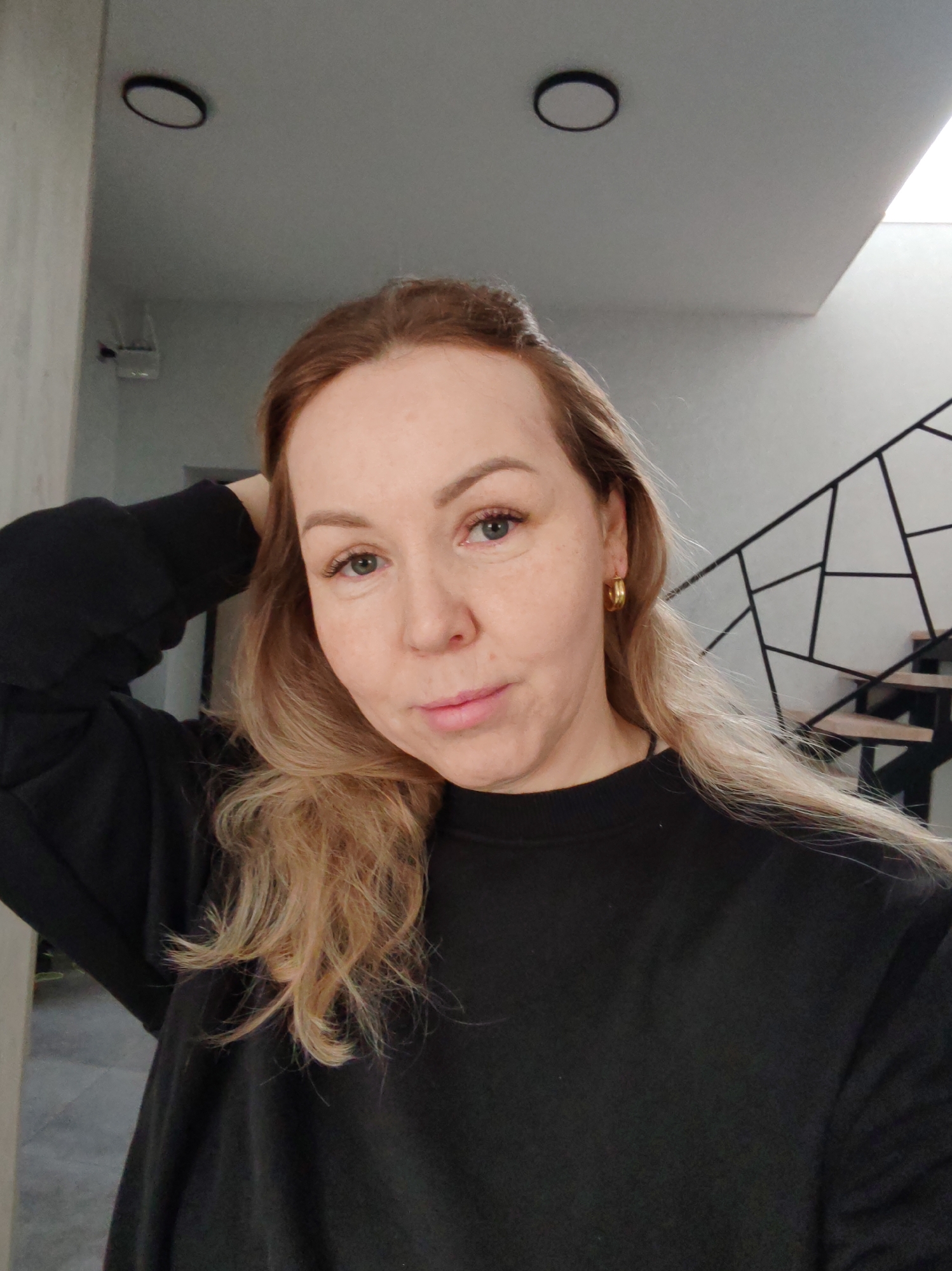 A woman is posing for a selfie indoors. She has light skin, blue eyes, and blonde hair. She's wearing a black sweatshirt and gold hoop earrings. Her left hand is raised to her head. In the background are two ceiling lights, a staircase with a geometric metal railing, and a skylight. There is no evidence in the image to confirm it was taken in Chaikovsky, Russia.