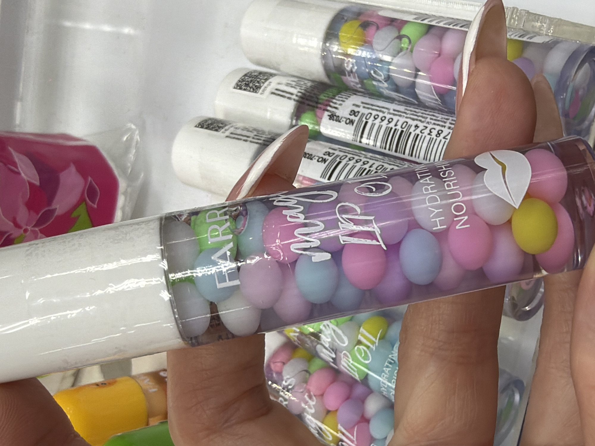 A human hand, featuring an almond-shaped fingernail with a white tip, holds a clear cylindrical tube of cosmetic lip oil. The tube, identified by text as "FARRI Magic LIP OIL - HYDRATING NOURISHING" and a white lip icon, contains numerous pastel-colored oval beads (pink, light blue, yellow, white) suspended in a clear liquid, and is sealed with an opaque white cap. Multiple identical or similar tubes are visible in the background, arranged on what appears to be a retail shelf or display. These background tubes feature white caps and barcode labels, one showing "NO.: 7038" and a number starting "783240166600". Additional blurry cosmetic products, including a yellow bottle and a pink item with a floral design, are present in the deeper background. The scene is located within a retail establishment in the city of Saint Petersburg, Russia.