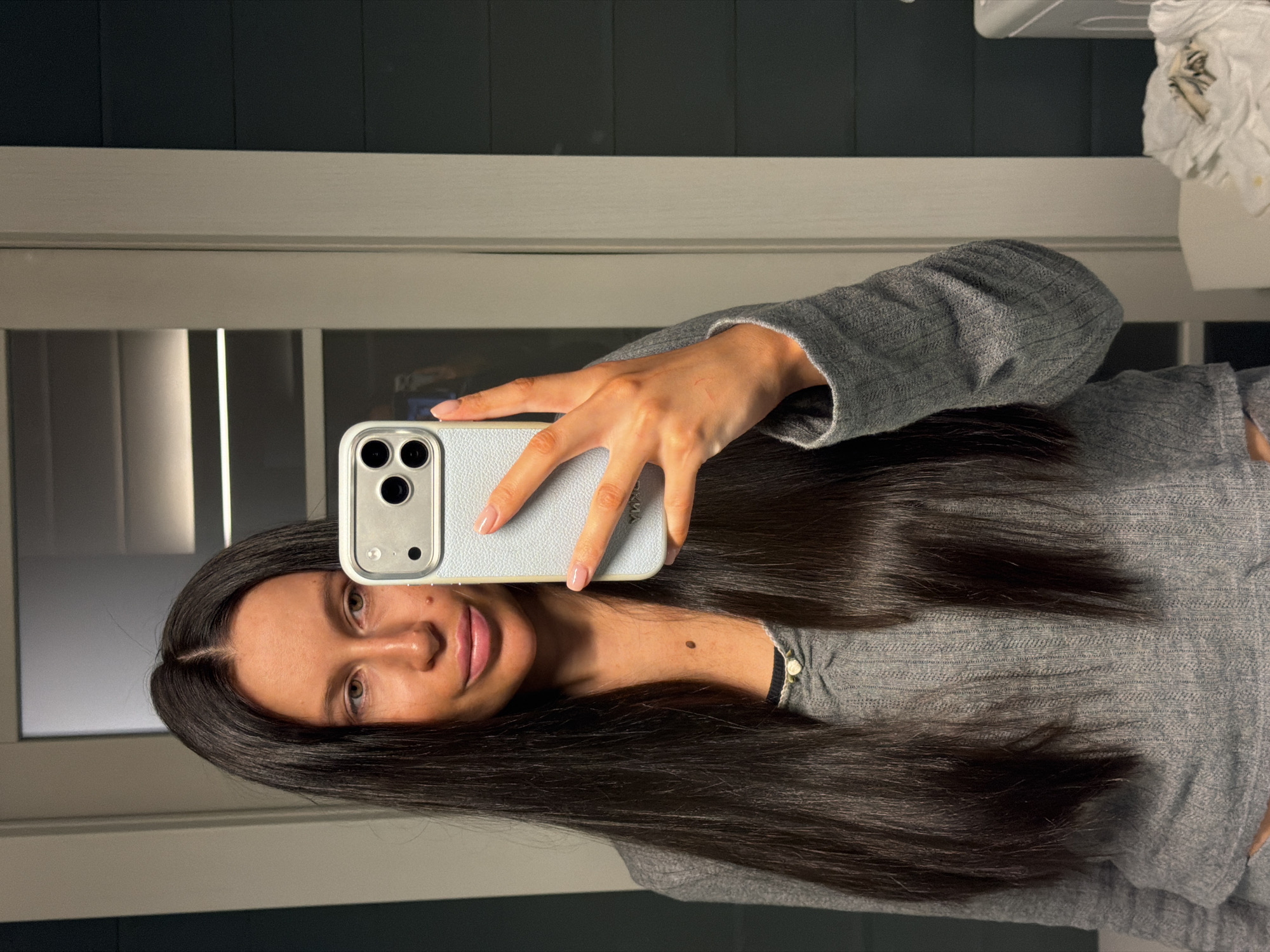The image displays a mirror selfie of a female individual with long, straight dark hair parted down the middle. She has a warm skin tone and her dark eyes are visible, with a neutral to slight smile on her lips. She is wearing a grey, ribbed long-sleeved top.
The subject is holding a light grey or silver smartphone, featuring a triple-lens camera, in front of her face, partially obscuring her upper left facial area. The phone is encased in a light-colored, possibly textured case. Due to the mirror reflection, reversed text is visible on the phone case, appearing to be "SYMA" or a similar word. Her fingers, with manicured nails, are visible on the phone.
The background suggests an indoor setting, likely a bathroom or changing room. Smooth, dark grey or black walls are visible above a lighter grey or off-white horizontal structure, which could be a shelf or trim. To the left, vertical lines hint at a door frame or window blinds. In the top right corner, a crumpled white item, possibly a towel or paper, is visible on a surface. The lighting is artificial, typical of an indoor space. There are no discernible elements indicating external weather conditions or a specific time of day. While the provided context suggests Khimki, Russia, no visual cues within the image confirm this location. The overall mood is casual and unassuming.