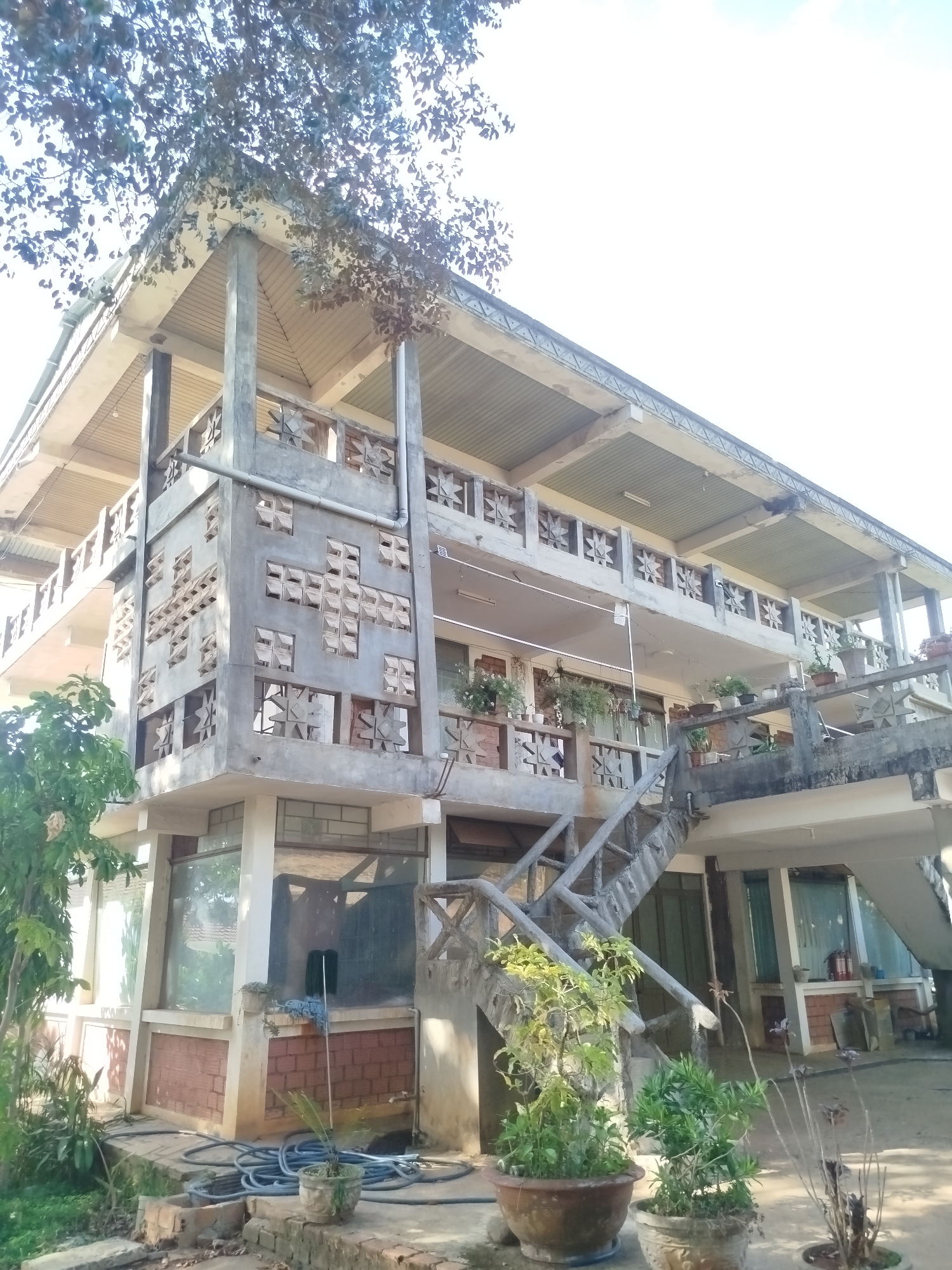 A two-story building stands in An Phu Ward, Vietnam. The building has a unique design with concrete walls featuring geometric cutouts. The lower level has glass windows and brick sections. A concrete staircase leads to the upper level, adorned with potted plants. The building is surrounded by foliage. A coiled blue hose lies on the ground near the base of the building.