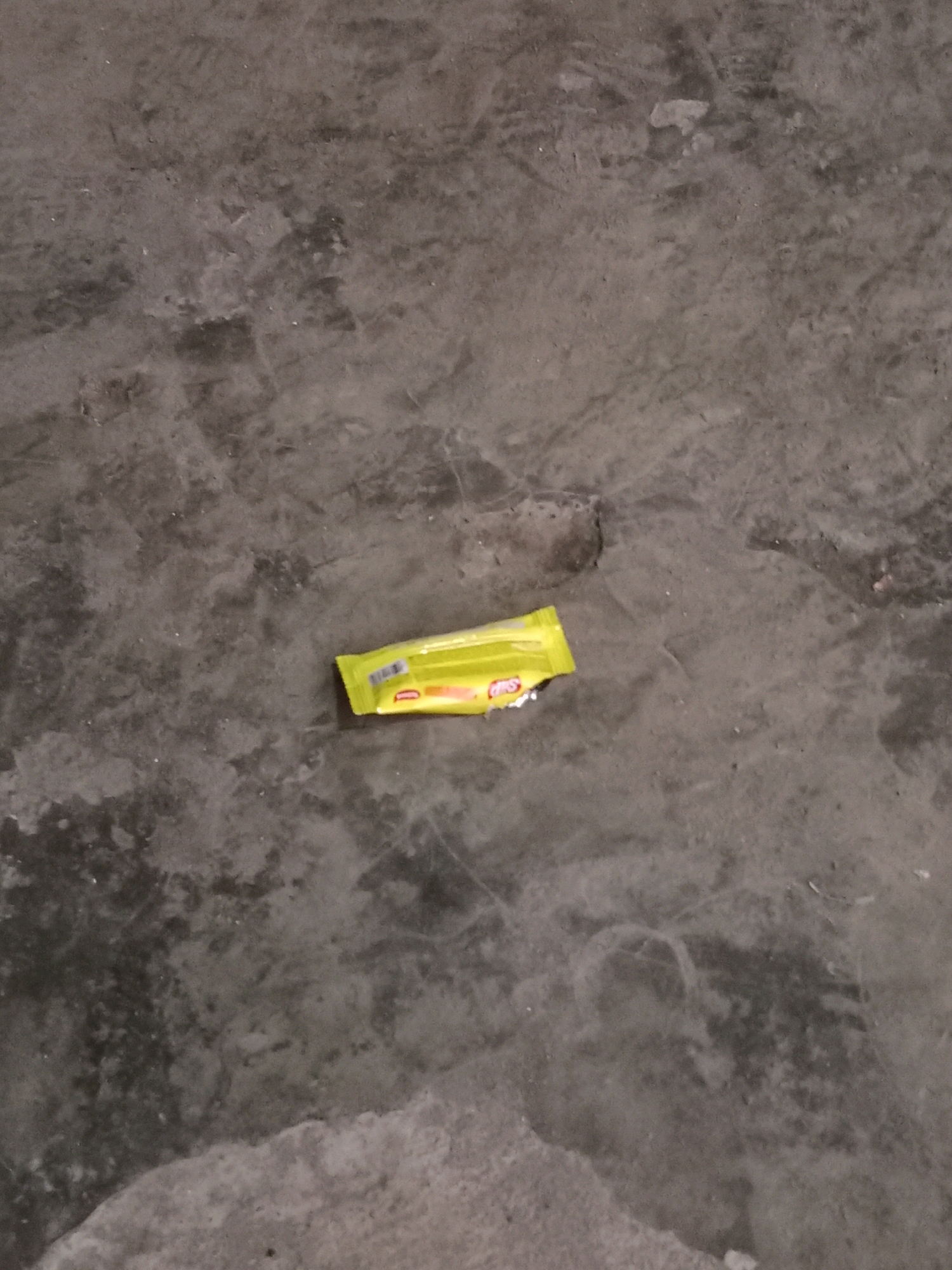 A single, unwrapped yellow candy wrapper is lying on a textured, gray concrete surface. The wrapper is oriented horizontally and appears to be slightly crinkled. The concrete surface shows variations in color and texture, suggesting it might be outdoors or in an unfinished interior space. There are no people, animals, or other objects visible in the immediate vicinity. The overall scene is mundane and appears to be a discarded piece of litter.