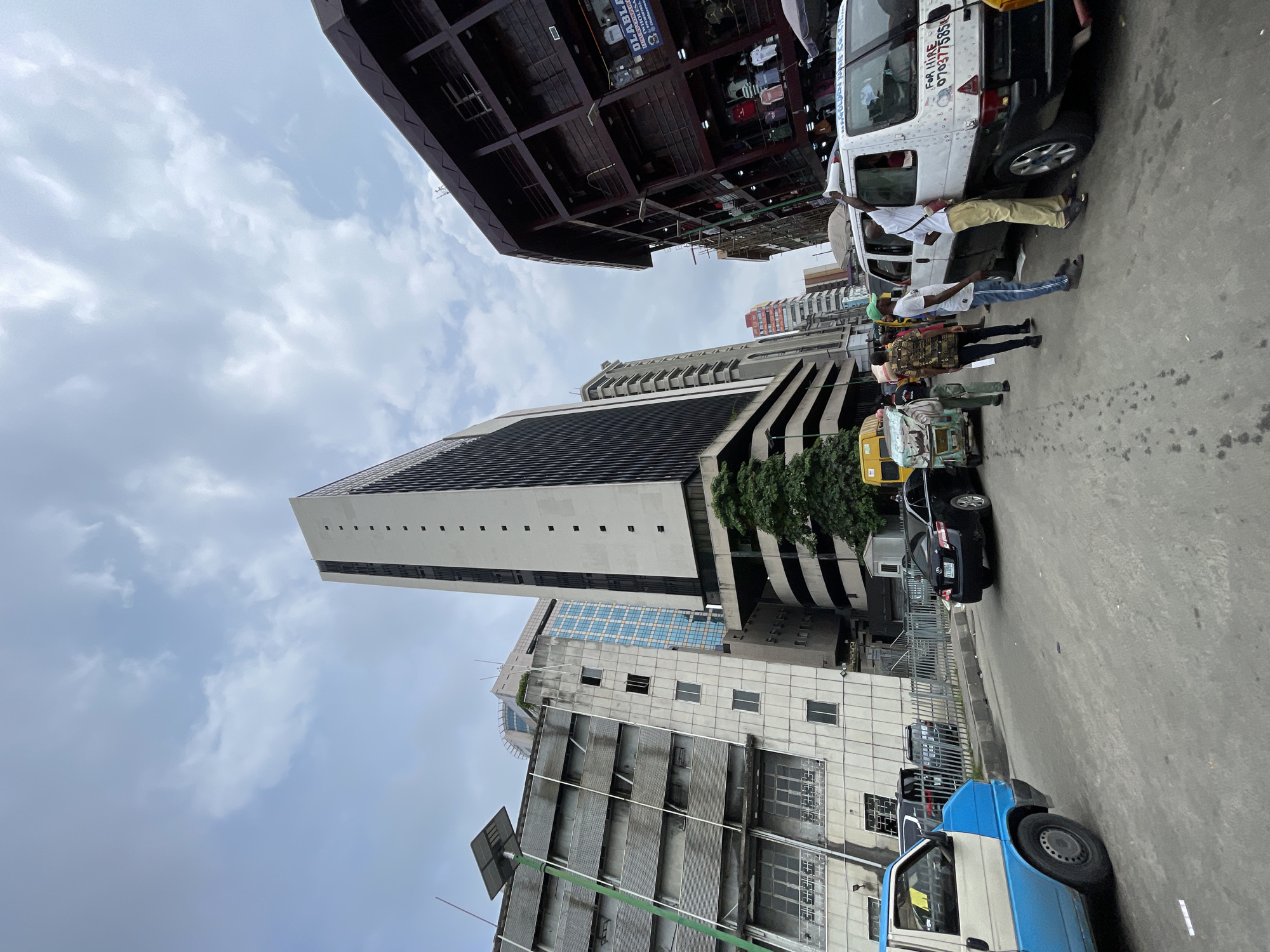 A low-angle, wide shot captures a street scene in Lagos, Nigeria, under a cloudy sky. The foreground is dominated by a section of a gray, textured asphalt road. To the left, the rear half of a blue and white van, with a gray wheel and visible wheel well, is parked. Next to it, a black sedan with a license plate reading "LXR 399 EK" is also parked. A chain-link fence runs along the sidewalk behind the vehicles.
Several people are visible on the sidewalk and street. A group of four men are gathered near a white van, one of whom, wearing a white t-shirt and khaki pants, is leaning into the van's open passenger window, holding a white object, possibly a megaphone. Another man in a green cap and blue jeans stands beside him, facing the van. Two other men, one in a patterned shirt and the other in a dark shirt, are also near the van. Further down the street, a person stands near a yellow and green rickshaw.
The background features a dense urban environment with several multi-story buildings. A prominent, tall, white building with black rectangular windows dominates the center of the frame. To its left, a gray building with horizontal windows and a solar panel on a green pole is visible. To the right of the tall white building, a dark, multi-story building with open-air levels displaying clothing racks is seen. In the distance, other buildings of varying heights and architectural styles are present. The overall impression is a bustling city street with a mix of modern and older structures, and significant human activity.