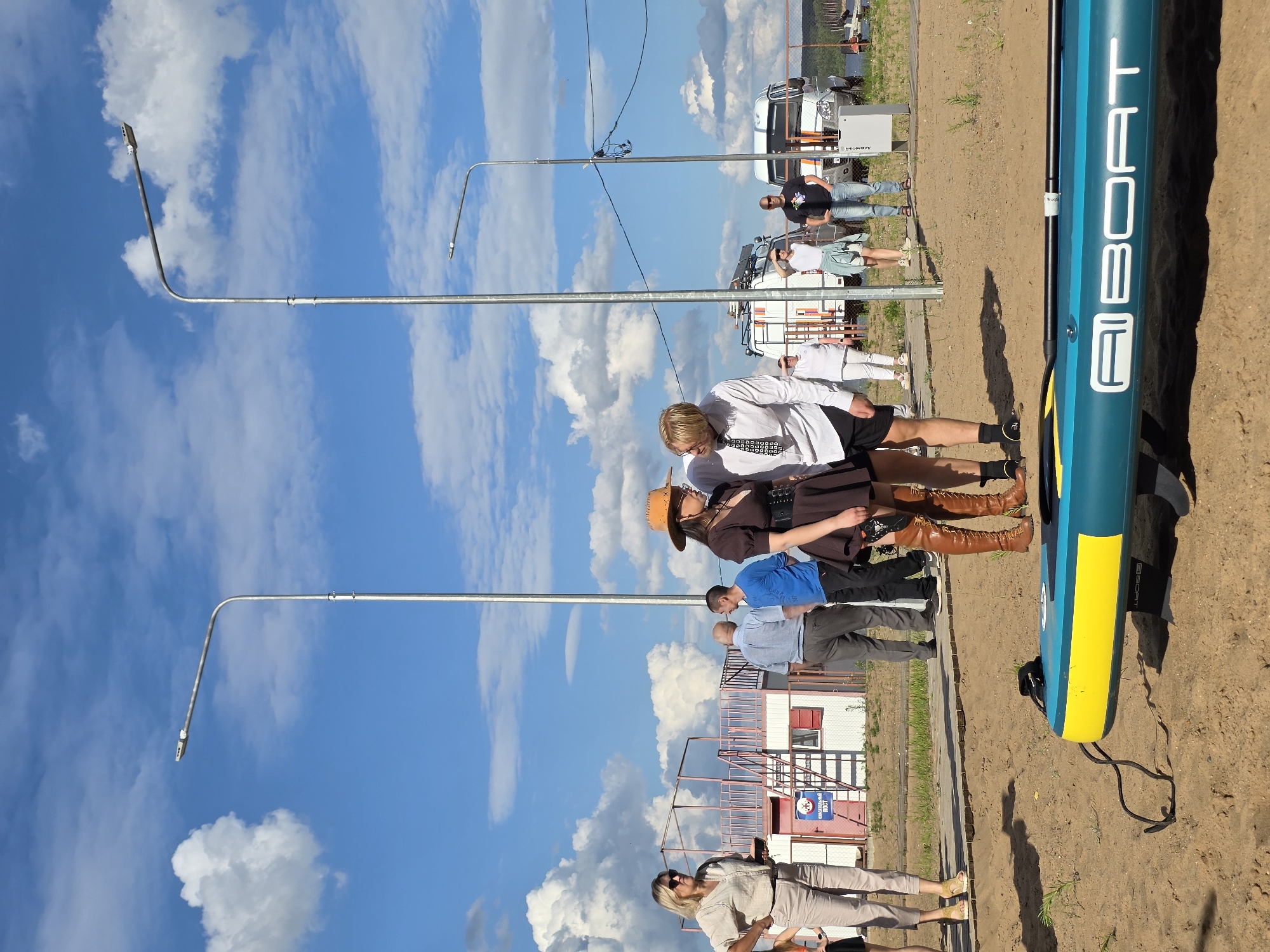 In Chaikovsky, Russia, a group of people stand on a sandy area near a body of water under a bright blue sky with scattered clouds. A teal and yellow paddleboard lies on the sand in the foreground. Two individuals, a man in a white shirt and tie and a woman in a brown dress and cowboy hat, stand close together, appearing to interact intimately. Other individuals are visible in the background, some near buildings and vehicles. One building is white with red trim and has stairs leading up to it. Two tall streetlights are prominent in the scene, with power lines connecting them. The overall atmosphere suggests an outdoor gathering or event.
