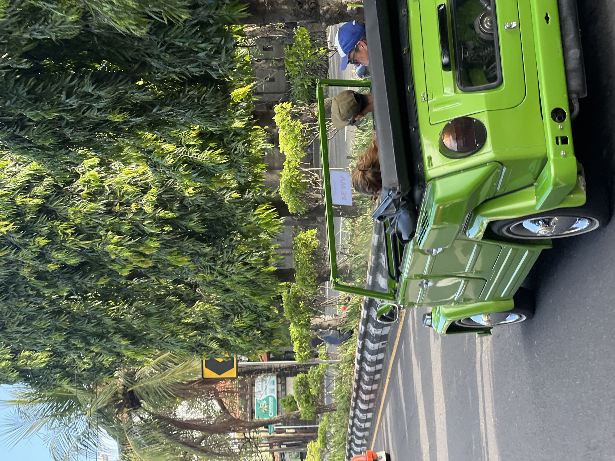 The image depicts a green, open-top vehicle with three occupants traveling along a road. The driver is wearing a blue baseball cap and glasses. The other two passengers are sitting behind him. The vehicle is on a road bordered by trees and shrubs on the left, and a painted curb with black and white stripes on the right. A black and yellow street sign is visible. The location appears to be Denpasar, Indonesia.