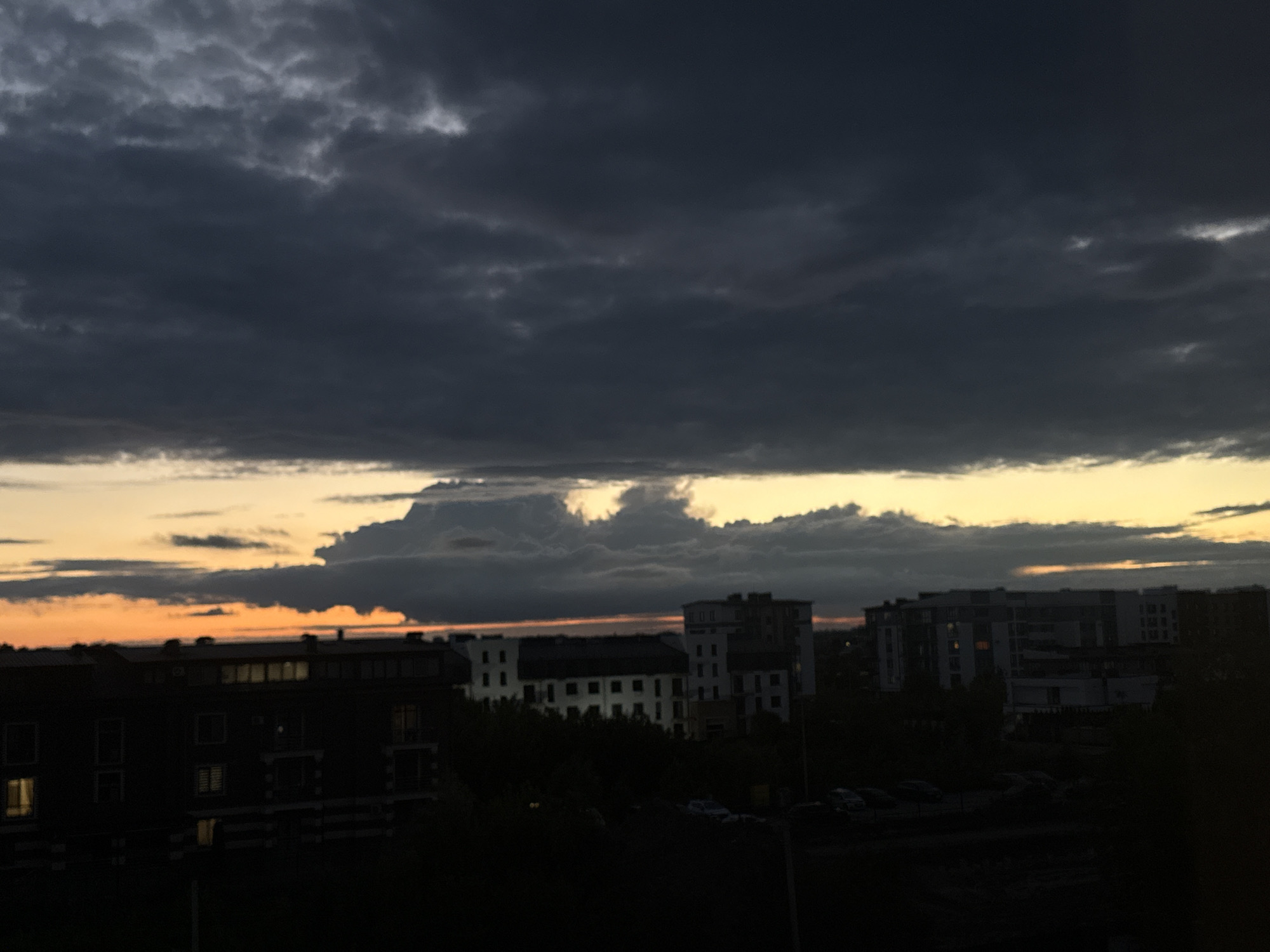 A dark, cloudy sky hangs over a cityscape at dusk. The clouds are thick and grey, with hints of yellow and orange peeking through the horizon, suggesting the last rays of sunlight. Below the sky, buildings of varying heights are silhouetted against the dim light. The closest building is a dark, multi-story structure with many windows, some of which are illuminated with a warm, interior light. Further in the distance, other buildings, some appearing to be apartment complexes, are visible. Trees and shadows obscure the ground level of the scene. Several cars are parked or driving in the lower portion of the image, mostly obscured by darkness. The overall impression is a moody, urban landscape transitioning into night.