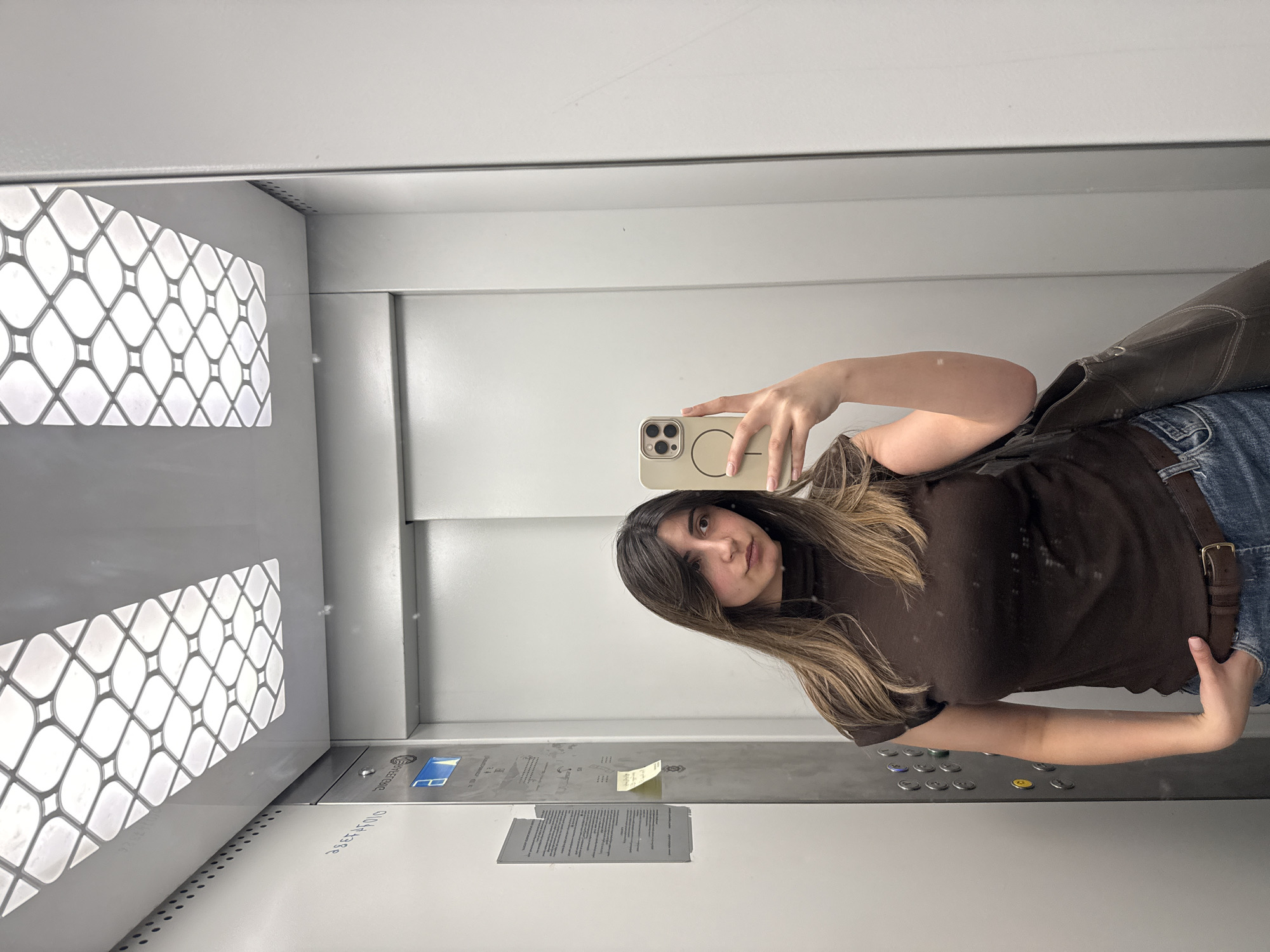 In an elevator, a young woman with long, dark brown hair and bangs takes a mirror selfie. She is wearing a dark brown, sleeveless turtleneck shirt and blue jeans with a brown belt. She is holding a beige iPhone in her right hand, and her left hand is in her jeans pocket. A dark brown shoulder bag is slung over her left arm. The elevator's interior is made of white metal panels, and the ceiling features a decorative light fixture with a patterned grid. A metallic panel with buttons and information signs is visible on the left wall. The scene is sterile and functional. The presence of a specific elevator model or signage could potentially indicate a location, but without further information or context, it is not possible to definitively place this image in Yerevan, Armenia, or any other specific city.