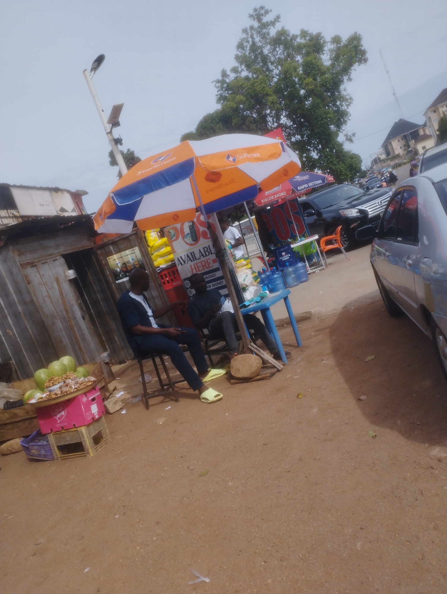 This is a medium shot, eye-level photograph taken outdoors in what appears to be a busy street market setting in Jos, Nigeria. The scene is dominated by a colorful umbrella providing shade for a vendor stall. The umbrella is predominantly orange and blue with "StarTimes" branding visible on it.
The stall itself is made of corrugated metal, suggesting a semi-permanent or temporary structure common in open-air markets. In the foreground, to the left, is a display of fruits, possibly guavas, on a tray atop pink and yellow crates. Next to the fruit display, a person is seated on a stool, facing away from the camera, wearing a dark blue traditional outfit and yellow sandals. They appear to be engaged in a conversation or interacting with someone out of frame. Further back, another person is seated on a stool behind a blue plastic table, also facing away from the camera, dressed in dark clothing.
Behind the stall, water dispensers and bottled water are visible, indicating that beverages are being sold. A black Lexus SUV is parked on the street behind the stall, along with other vehicles and a few people in the background, suggesting a relatively active street with vehicular traffic and pedestrian presence. The ground is covered in reddish-brown dirt.
Visible text on the umbrella includes "StarTimes" and "YIELDS MORE RAPID SETTING". On a sign behind the stall, "POS", "AVAILABLE HERE", "MONEY", and "AW MONEY" are partially legible. The lighting suggests it might be daytime, possibly overcast or late afternoon, given the diffused light and absence of strong shadows. The overall impression is a typical street-side vending operation in a bustling Nigerian urban environment.