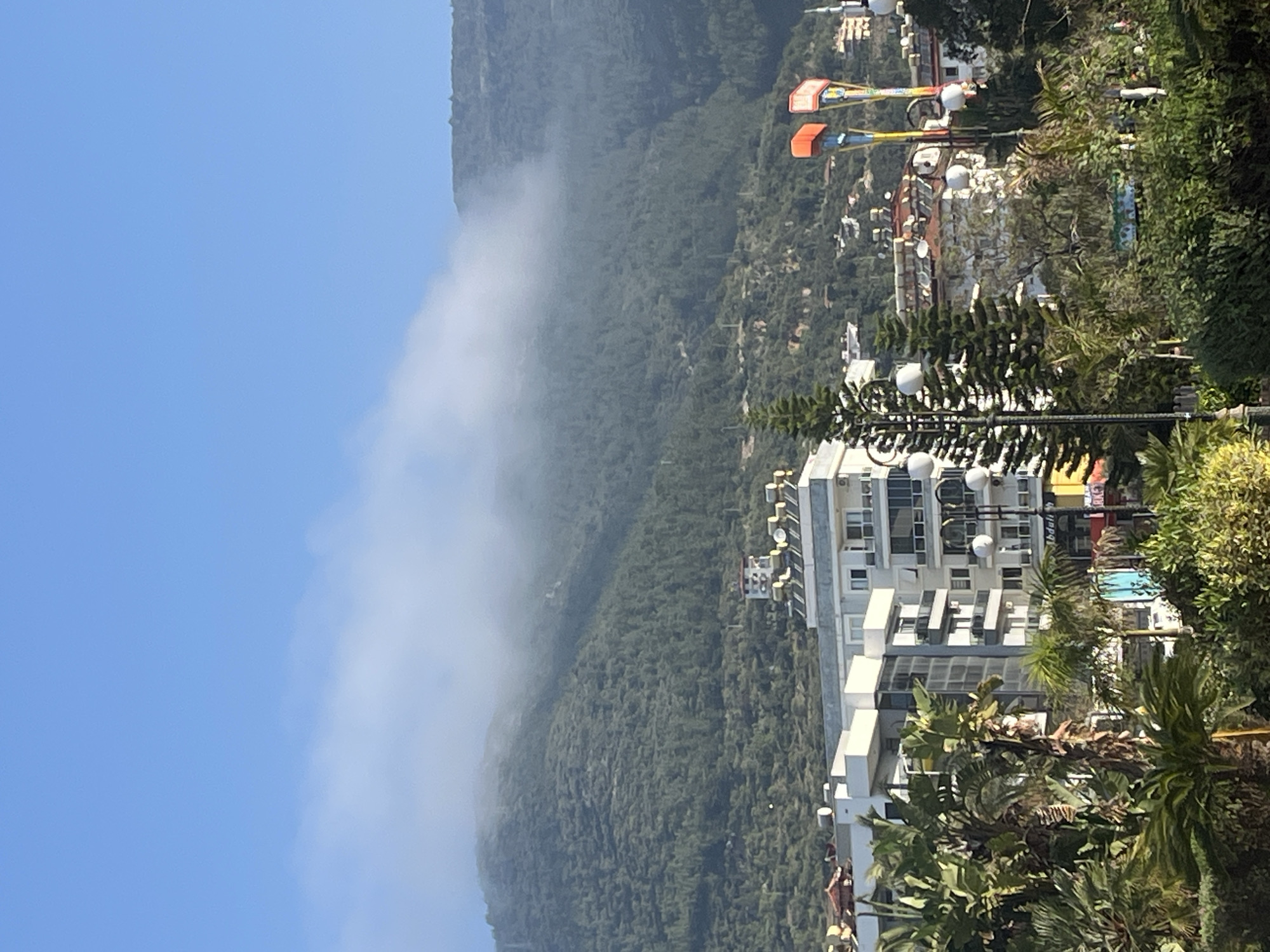This media file captures a bright, daytime scene of a town situated at the base of a large, green, forested mountain range. The image appears to be taken from an elevated perspective, possibly from a building or a hillside overlooking the area.
In the background, a vast, densely wooded mountain dominates the left side of the frame, its upper slopes partially obscured by a long, horizontal band of white cloud or mist that clings to the mountainside. Above the cloud line, the sky is a clear, vibrant blue.
The mid-ground and foreground feature a collection of modern, multi-story buildings, predominantly white or light-colored, with numerous windows and balconies. These structures suggest hotels or residential complexes. Among the buildings and lush green foliage, several prominent palm trees and other tropical-looking plants are visible, adding to the warm climate aesthetic. Two distinct, tall structures with bright yellow supports and orange, basket-like seats are visible on the right side, resembling amusement park rides, possibly a slingshot or similar thrill ride. Streetlights with multiple spherical white lamps are also scattered throughout the scene.
The time of day is bright and sunny, indicated by the clear blue sky and strong illumination across the landscape. The weather appears fair, though with the notable mist on the mountain. There are no people visible in the image, suggesting a tranquil moment. No clearly decipherable text is visible on any of the buildings or signs, although some faint, unreadable characters might be present on distant signs.
Given the architectural style, dense vegetation, and the presence of resort-like elements, the scene is consistent with a popular tourist destination, specifically Alanya, Turkey, as provided by the location context. The overall impression is one of a scenic and pleasant urban landscape nestled against a dramatic natural backdrop.