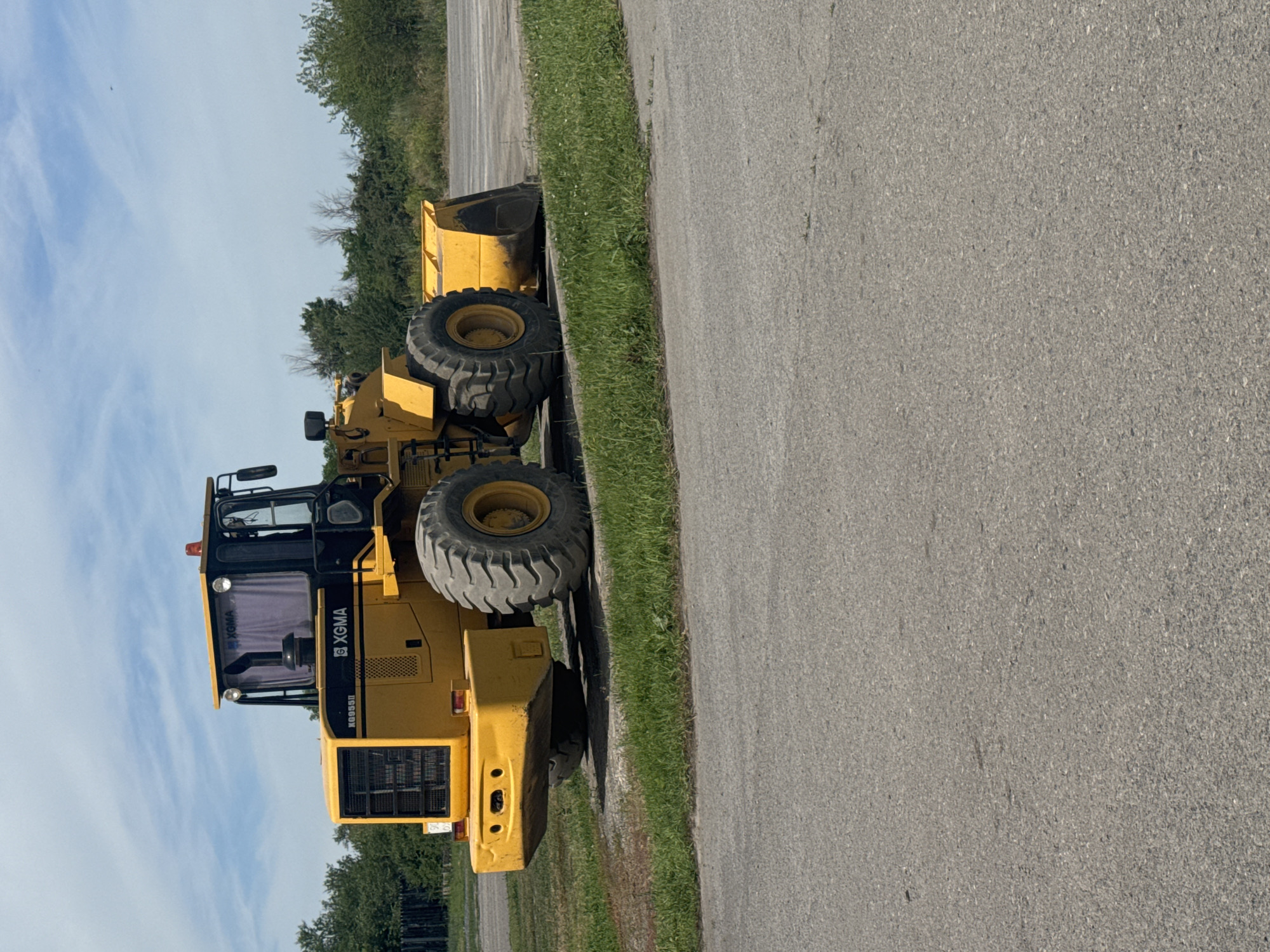 A yellow XGMA wheel loader is parked on an asphalt surface next to a strip of grass. The wheel loader is facing right and is viewed from the front-right. The machine has large, treaded tires, a raised cab with tinted windows, and a bucket at the front. The background features a line of green trees and a cloudy blue sky. The scene appears to be an outdoor industrial or construction area. The location is in Shakhty City District, Russia.