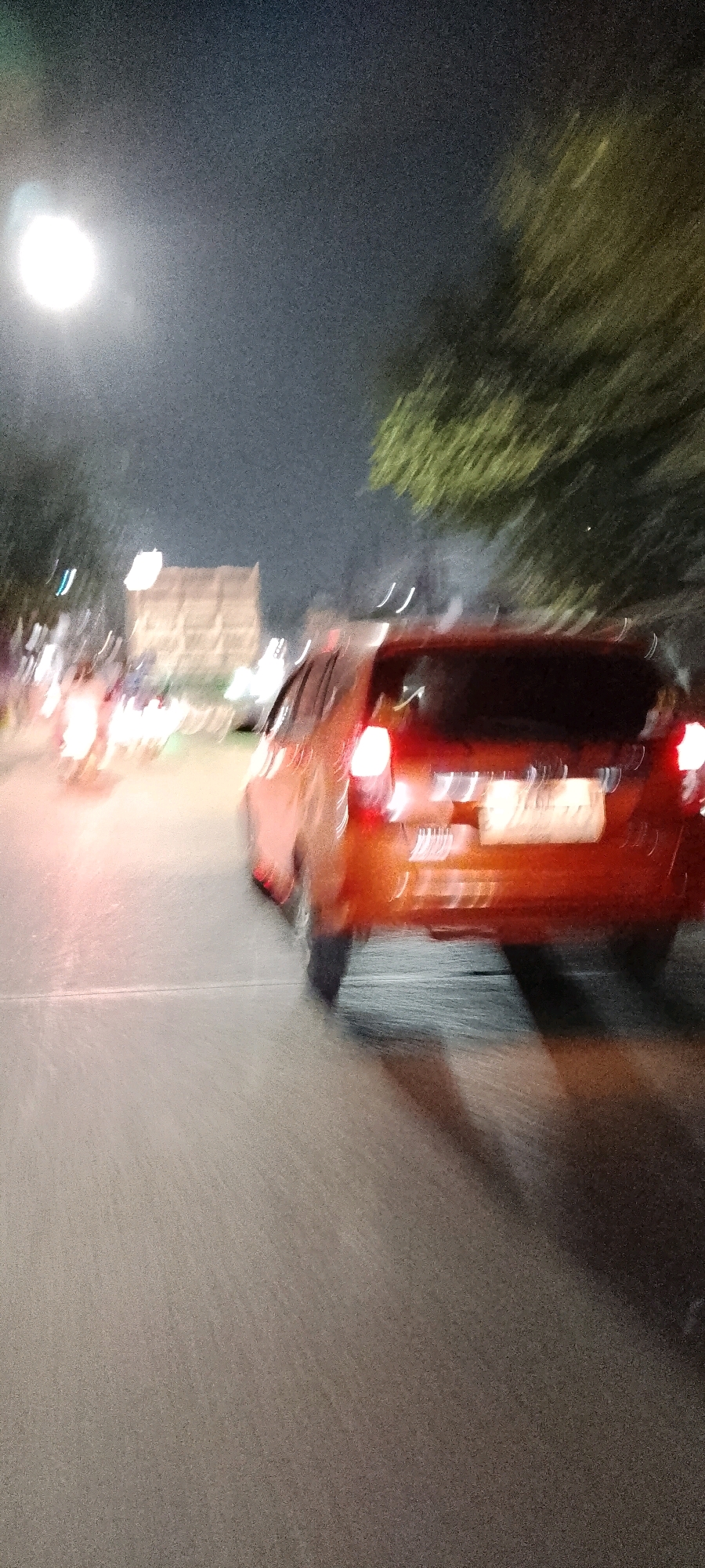 The image depicts a motion-blurred nighttime scene on a city street in Cileungsi, Indonesia. In the foreground, the rear of an orange car is prominently visible, its taillights illuminated. The car appears to be in motion, as indicated by the streaking lights. To the left of the car, there are blurred streaks of light suggesting other vehicles, possibly motorcycles. In the background, a building with illuminated windows is visible, partially obscured by trees with blurred foliage. The overall environment is dark, illuminated by artificial lights and the headlights/taillights of vehicles. The ground is a paved road.