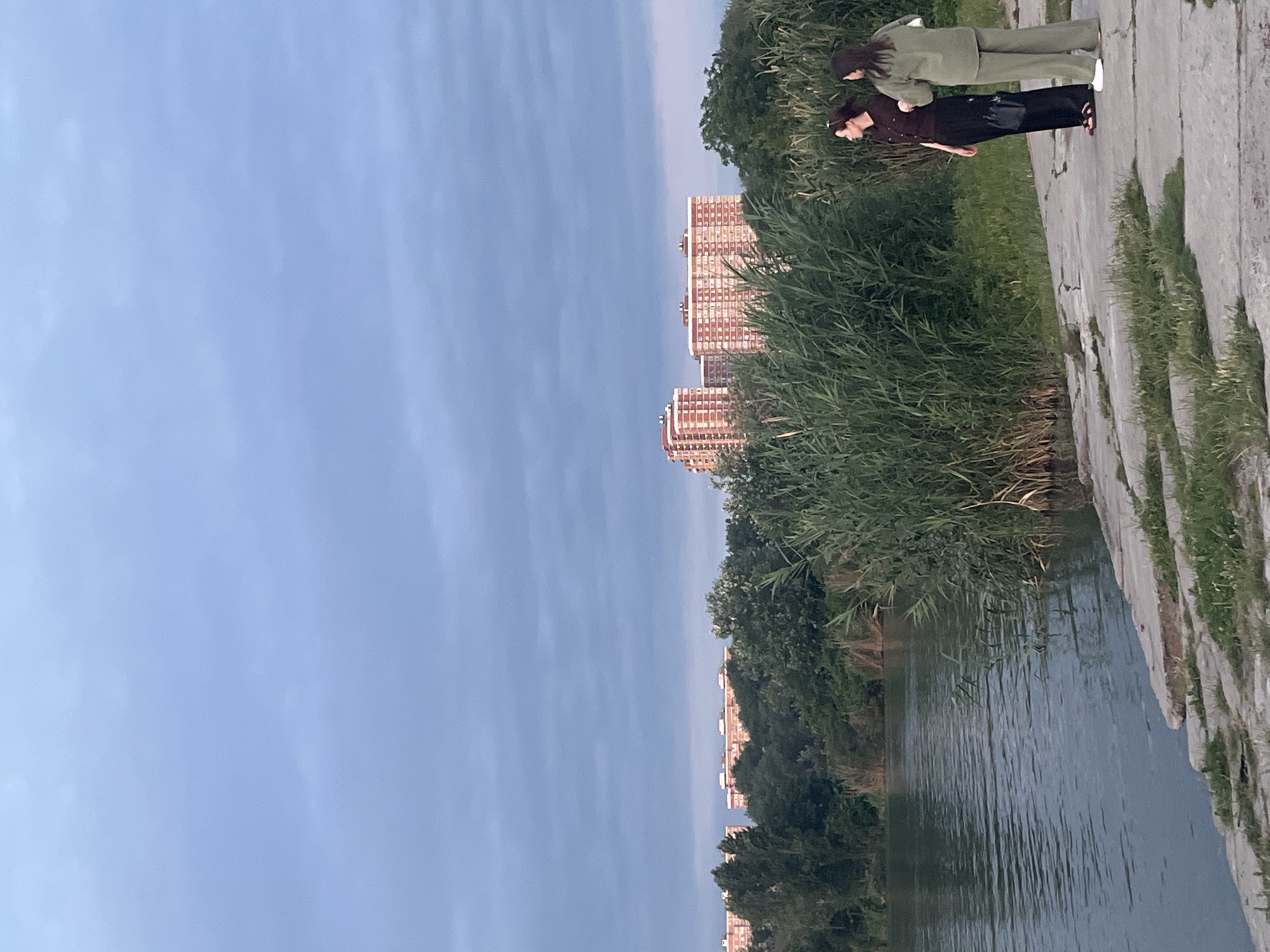 Two women stand on a concrete embankment overlooking a body of water. The woman on the left wears a dark maroon short-sleeved shirt and black pants. The woman on the right wears a light green outfit consisting of a long-sleeved top and loose pants. They are facing away from the camera, looking towards the water and the vegetation on the opposite bank. Behind the vegetation, several multi-story apartment buildings are visible, their facades predominantly white with reddish-orange window frames and accents. The sky above is overcast with a pale blue hue. The scene appears to be a park or recreational area near the water, possibly the Kuban River in Krasnodar, Russia, given the architectural style of the buildings and the presence of dense greenery. The lighting suggests it is either early morning or late evening.