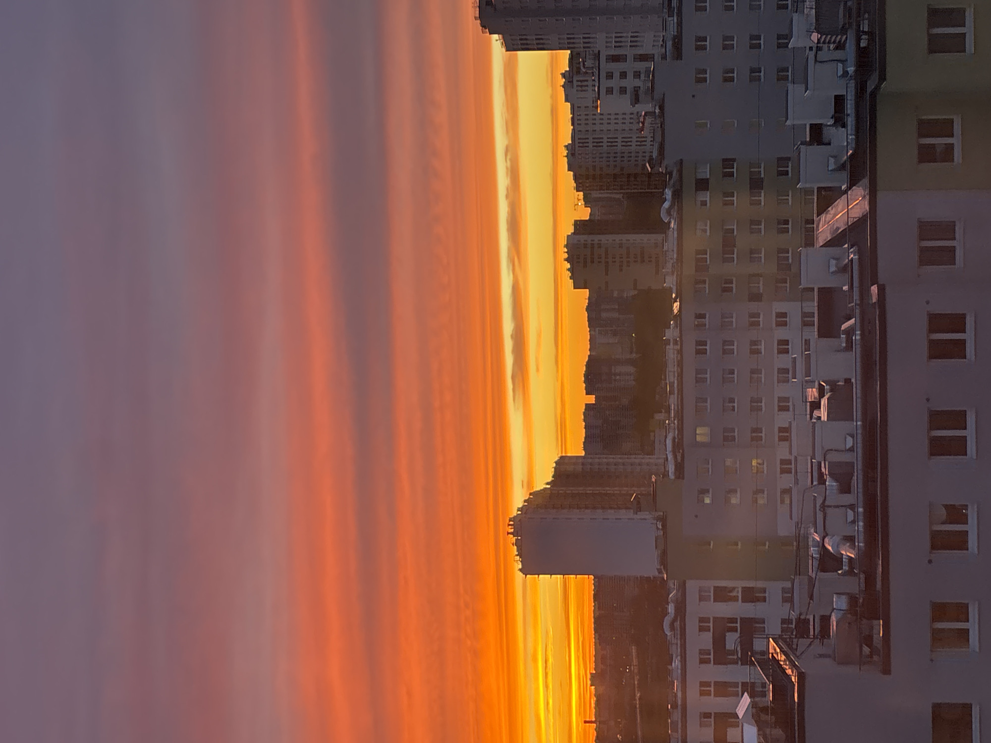 The image shows a sunset over the city of Vsevolozhsk, Russia. The sky is a gradient of orange and purple, with the sun illuminating the clouds with a bright yellow glow. In the foreground, there are apartment buildings. The buildings are mostly white and gray, with some yellow and green accents. There are many windows visible on the buildings.