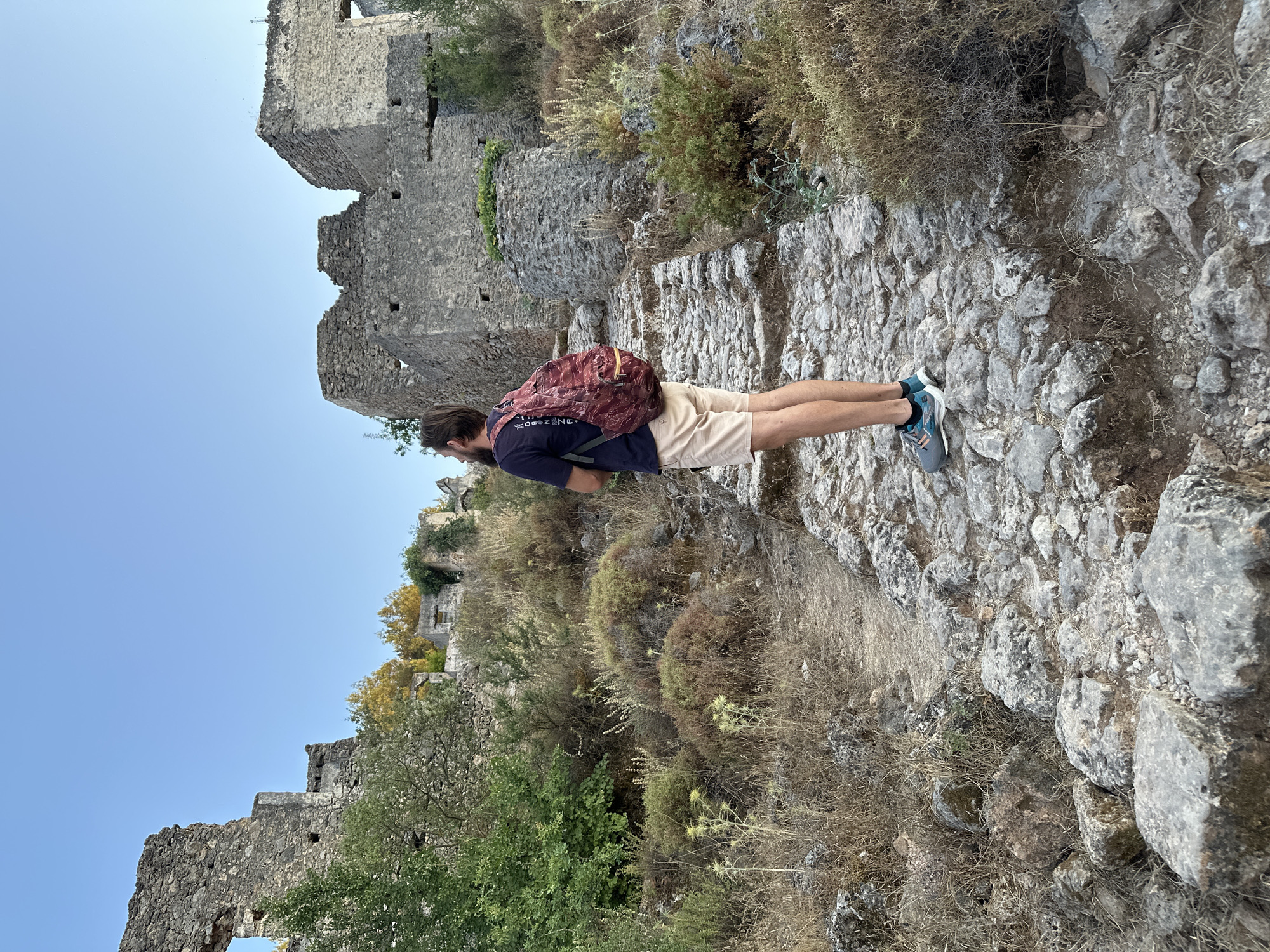 A person with a beard wearing a dark blue t-shirt, beige shorts, and athletic shoes stands on a cobblestone path, looking towards ancient stone ruins under a clear blue sky. The person has a maroon and gold patterned backpack. The ruins, which appear to be old stone structures, are overgrown with vegetation. The environment is dry and arid, with sparse grasses and shrubs. The scene suggests an archaeological site or historical landmark, likely Kayaköy, a Greek ghost village located near Fethiye, Turkey, known for its well-preserved ruins.