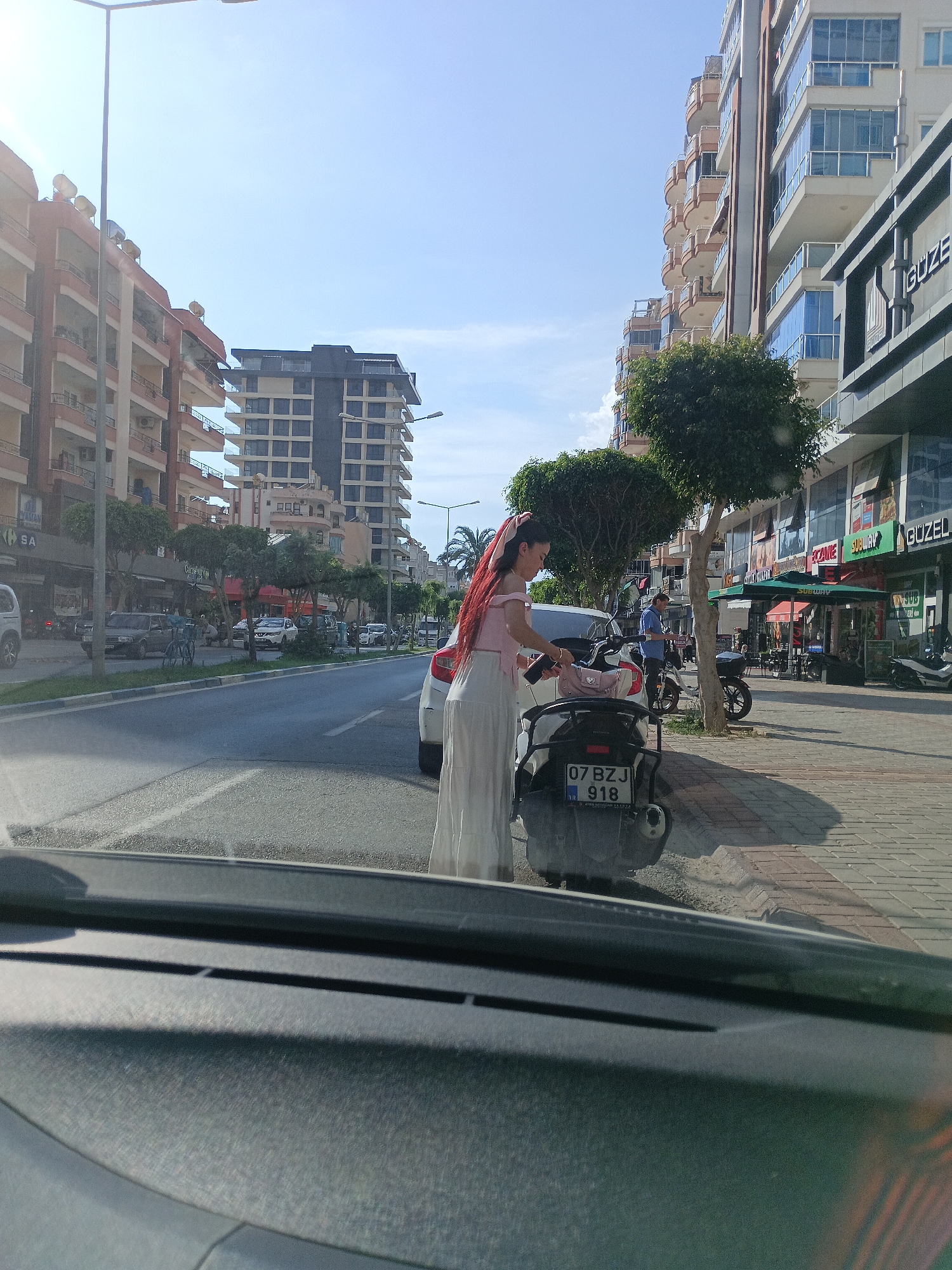 The image depicts a street scene in Alanya, Turkey, likely during daylight hours given the bright sky. In the foreground, the interior of a vehicle is visible, showing a dashboard and windshield. The perspective is looking out towards a street lined with multi-story buildings on both sides. On the right side of the street, there are shops, including one with "SUBWAY" signage and another with "GUZE." Several trees are planted along the sidewalk.
In the middle ground, a young woman with long, red hair is standing next to a white and black scooter. She is wearing a pink off-the-shoulder top and a long, light-colored skirt. She appears to be holding a phone and a pink bag. A man in a blue shirt is standing near a parked motorcycle a short distance away. Several cars and scooters are parked or driving on the street. The buildings are modern, with balconies and large windows. The street is paved with asphalt, and the sidewalk is made of brick pavers. The license plate on the scooter is clearly visible and reads "07 BZJ 918." The overall impression is a bustling urban environment.