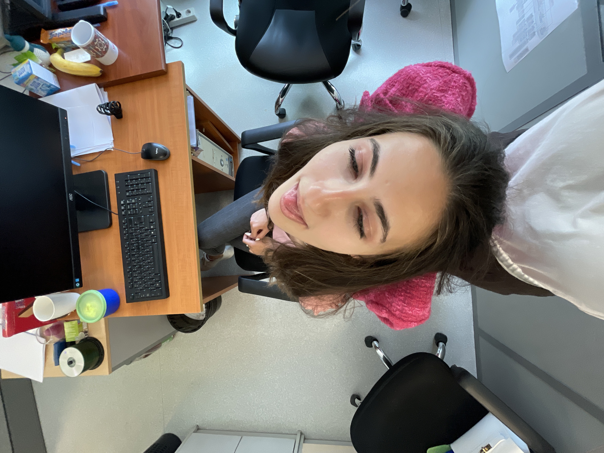 This high-angle, notably rotated image captures a young woman seated at a brown wooden desk in an indoor office or study setting in Sofia, Bulgaria. She has long, dark brown hair and fair skin, and is wearing a pink textured top and dark pants. Her eyes are closed, and her lips are slightly parted, revealing her tongue, conveying a relaxed and playful expression. A white fabric is draped over her right shoulder.
The desk is populated with various items. A black computer monitor (with "AOC" visible) and a black keyboard are positioned in front of her. To the right of the keyboard is a black Logitech computer mouse. Other objects include a white ceramic mug, a banana, and a blue box with "MILK" partially visible. A chocolate bar wrapper, with "To...Pobar" or "Topob...ar" visible, sits nearby. Several papers are held by a black binder clip, and a blue pen is also on the desk. Towards the lower left of the desk, a blue and green lidded container, a roll of tape, and other small supplies are present.
The background reveals two black office chairs with chrome bases, and a light-colored floor. A power strip with plugs is visible near the top right of the desk. A document is posted on a wall further back. The scene suggests a casual, candid moment during a break in an active workspace.