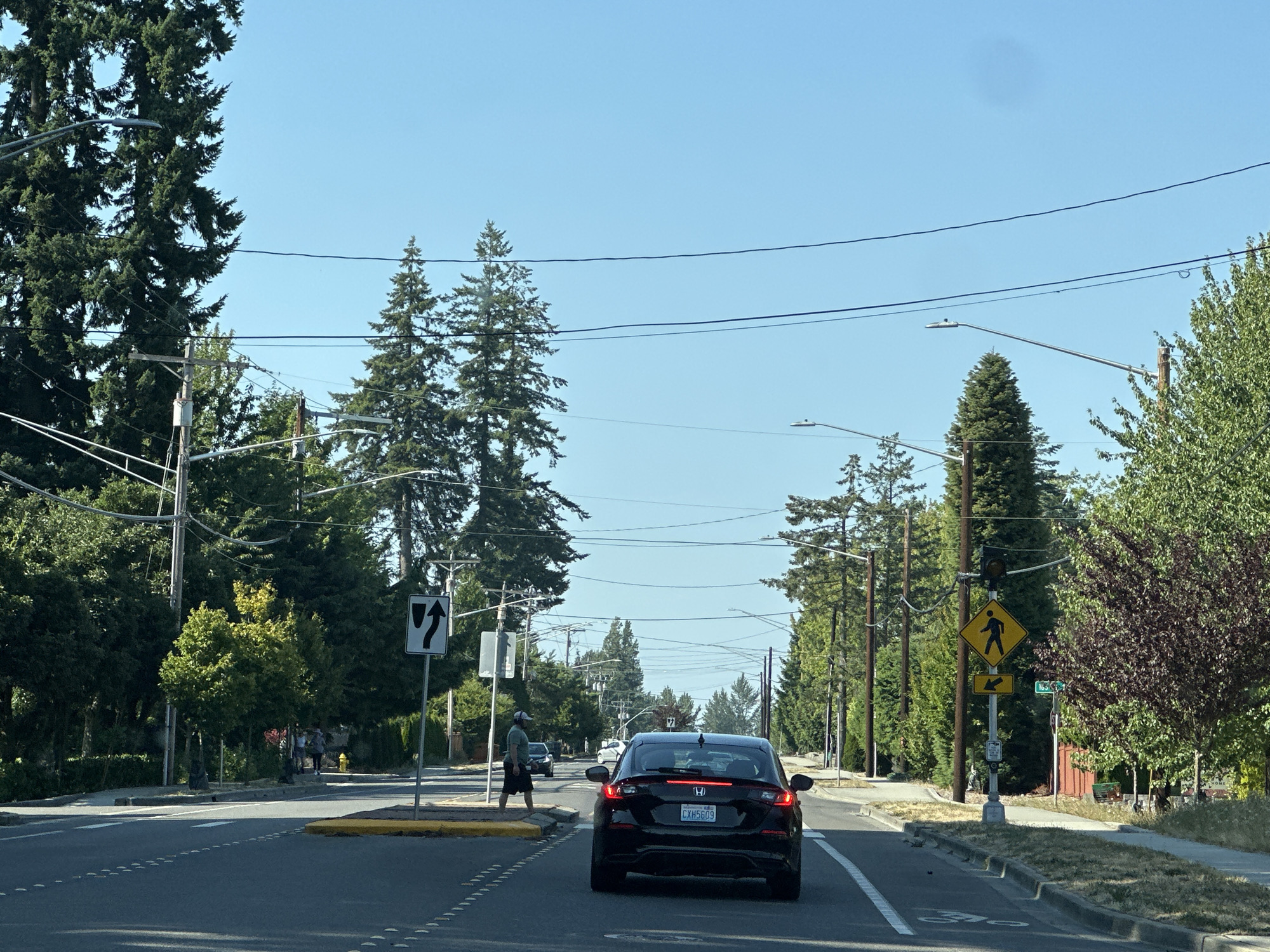 This is a real-world image taken from a vehicle traveling on 164th Street Southwest in Lynnwood, Washington, United States of America. The scene depicts a residential street with mature trees lining both sides of the road. A black Honda hatchback is in the foreground, with its rear clearly visible. Several other vehicles are visible further down the road, along with a pedestrian crossing the street. Traffic signals and street signs are present, including a pedestrian crossing sign and a sign indicating a lane split. The sky is clear and blue, suggesting a sunny day. The overall impression is one of a suburban environment during daylight hours.