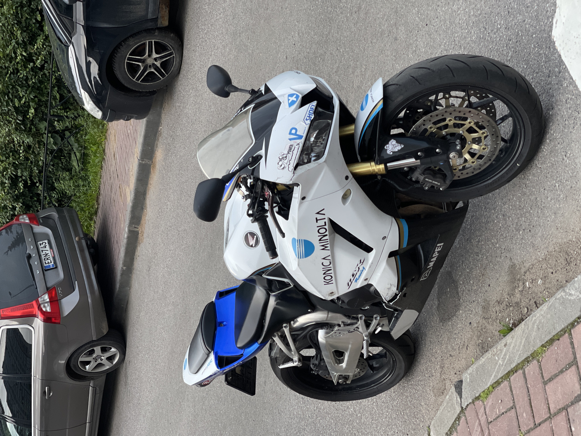 In the city of Unknown, Unknown, a white and blue sport motorcycle is parked on an asphalt street. The motorcycle has a black seat and a blue tail section. Various sponsor decals are visible on the motorcycle, including "KONICA MINOLTA" and "MAPEI". The motorcycle is positioned in front of a grey Volvo V70 station wagon and a black car, both parked along the curb. A brick sidewalk runs alongside the street, bordered by a concrete curb. Green foliage is present behind a black metal fence.