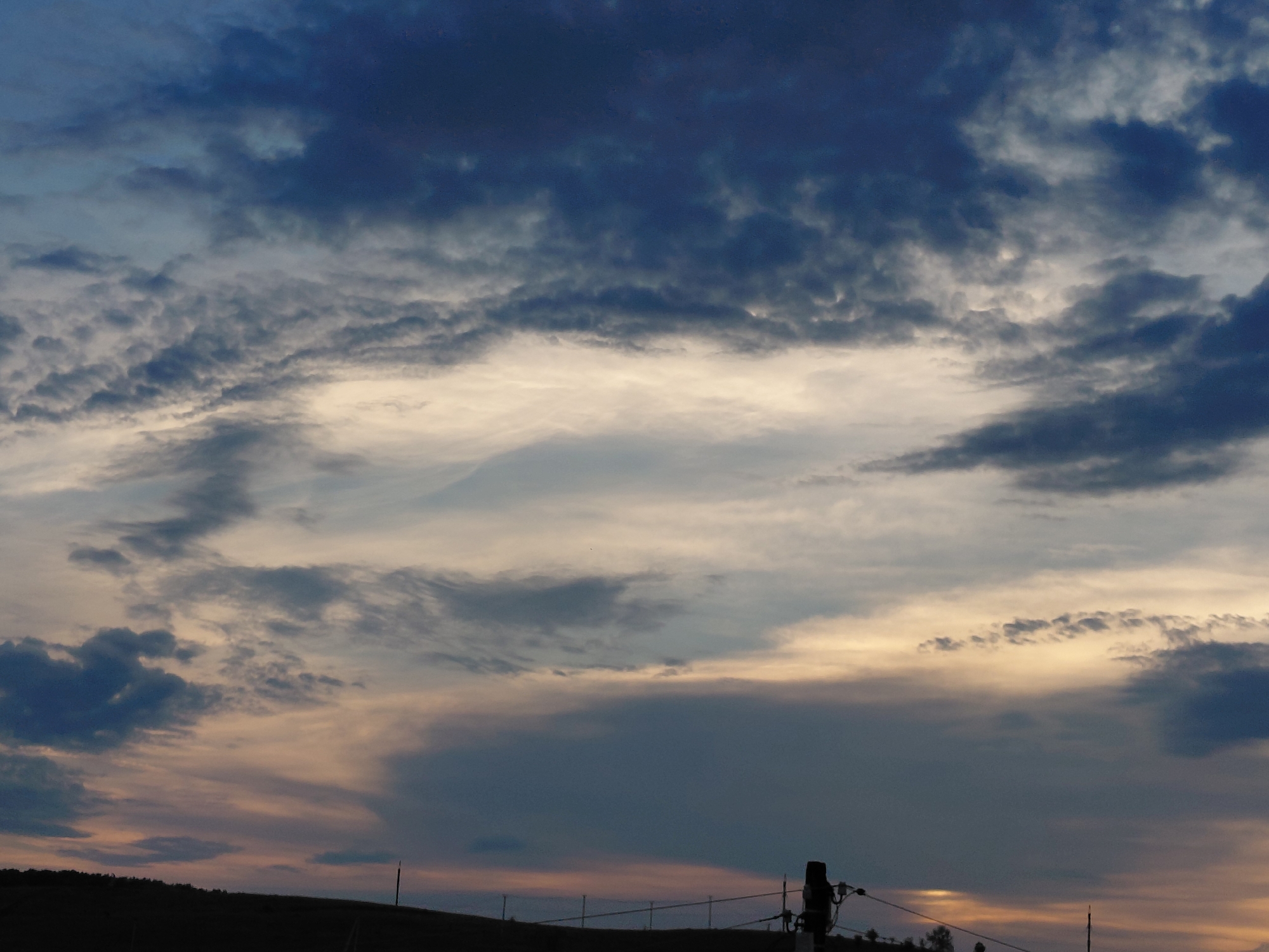 This is a photograph of a cloudy sky over a silhouetted landscape, likely taken at dusk or dawn. The sky is filled with a mix of dark blue, grey, and lighter, warmer tones, suggesting the presence of both storm clouds and the fading light of the sun. The clouds are varied in texture, with some appearing wispy and others more dense.
In the foreground, a dark, hilly silhouette is visible across the bottom of the frame. A few thin, horizontal lines, possibly fences or wires, stretch across this silhouetted area. To the right, a dark, vertical object, possibly a utility pole with some attached equipment, is prominent. Another smaller vertical element is visible further to the right. The overall impression is of a natural landscape under a dramatic sky. This scene could be found in or near Novokuybyshevsk, Russia, a city known for its industrial areas and surrounding natural landscapes. The image captures a specific moment in time, emphasizing atmospheric conditions and the transition of light.