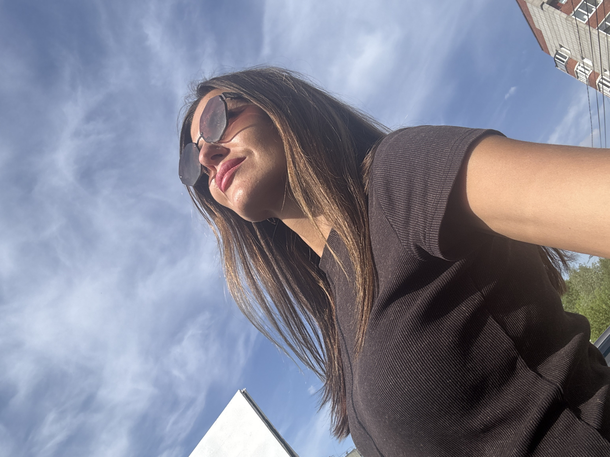 A low-angle shot depicts a woman in sunglasses against a blue sky with wispy clouds. She is wearing a dark, ribbed shirt. In the background, the corner of a white building is visible in the lower left, and the corner of a taller building with multiple windows is in the upper right. This location is in the city of Chaikovsky, Russia.