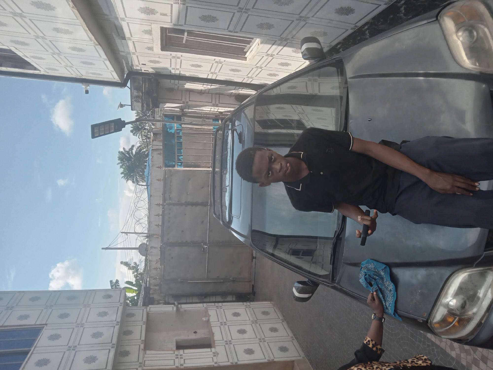 A young man in a black polo shirt and dark pants is lying on the hood of a black car. He is holding a black object in his right hand. An individual wearing a leopard print garment is partially visible in the lower right corner, holding a blue patterned cloth on the car's hood. The car is parked outside a building with tile-patterned walls and windows. In the background, there is a metal gate topped with barbed wire, along with utility poles and a street light. The sky is visible with some clouds. The location is the city of Owa-Alero, Nigeria.
