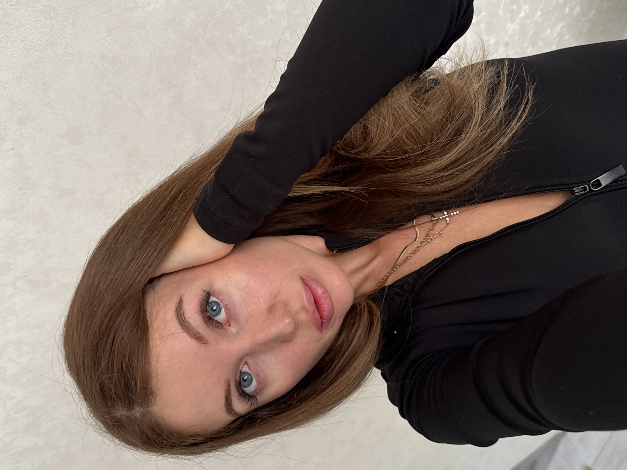A woman with long brown hair and blue eyes looks directly at the camera. She is wearing a black, long-sleeved top with a zipper, and a gold necklace with a cross pendant. Her hand is resting on her head. The background appears to be a plain, light-colored wall. It is not possible to identify the location as Chaikovsky, Russia from this image alone.